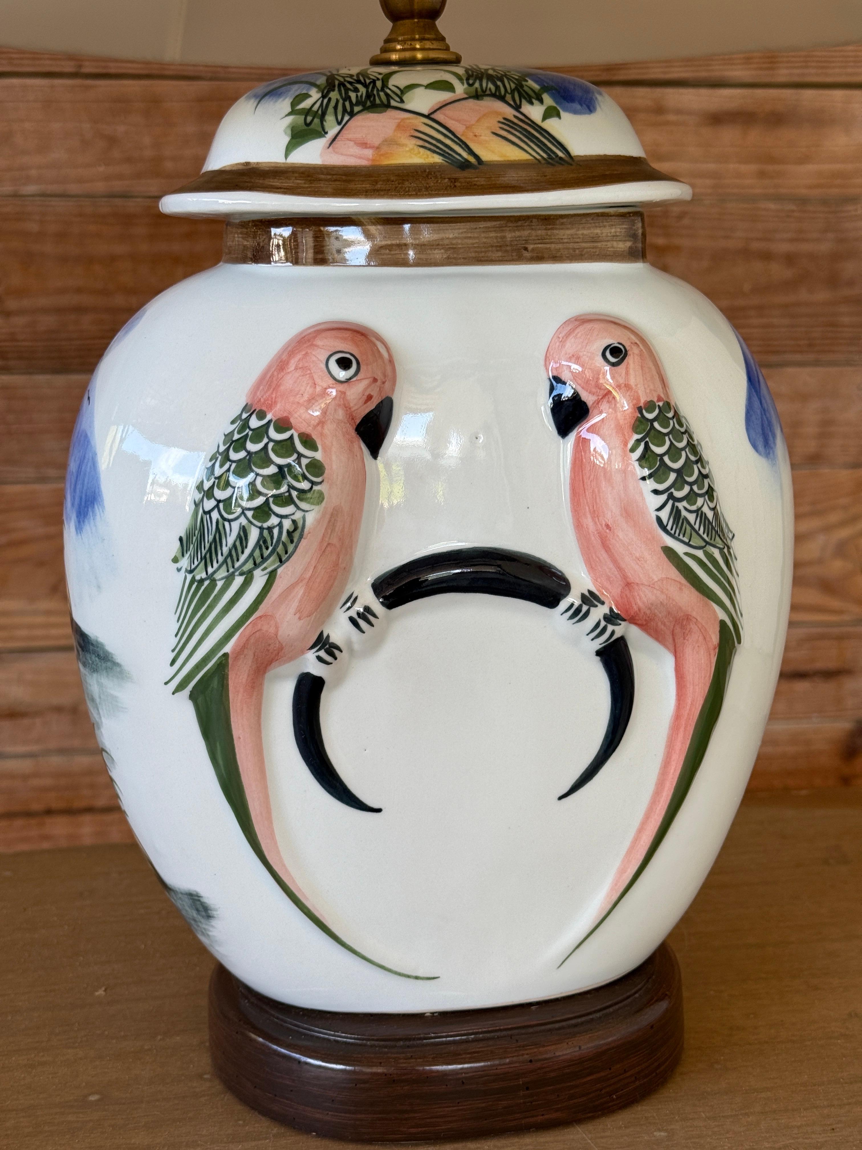 Une vibrante lampe vintage en céramique peinte à la main avec de charmants motifs de perroquets en rose corail et vert, perchés sur un perchoir circulaire. Le verso présente un paysage coloré avec des montagnes, des arbres et des oiseaux en vol.