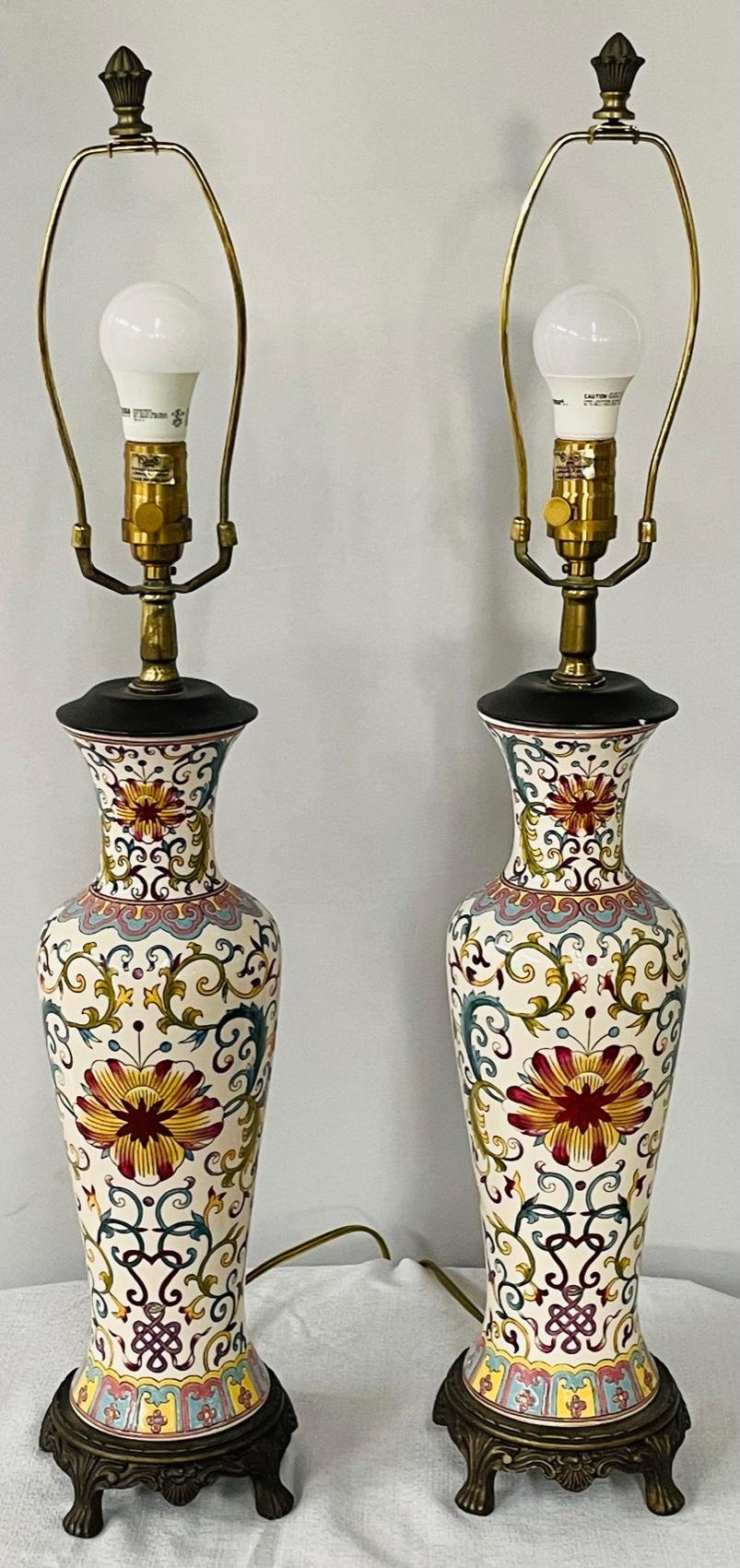 A 1970's pair of hand painted porcelain lamps in a shape of urns featuring a multi-colored geometrical motif design and raised on a wooden base. The lamps comes with Boho chic style linen shades decorated with small black tassels. Very chic, the