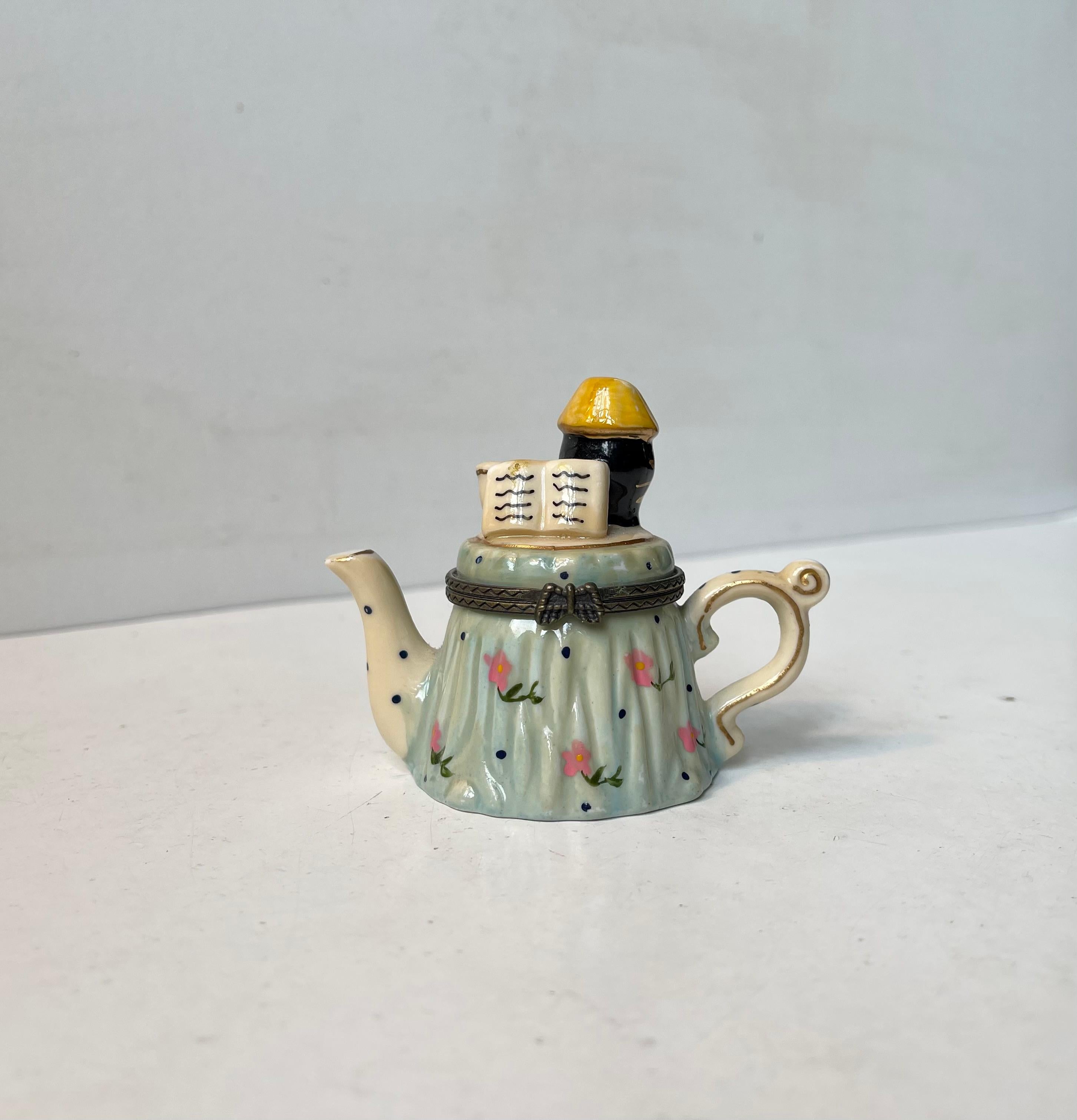Curios novelty trinket in the shape of a porcelain teapot trinket hand-painted with pink flowers, a coffee cup and a hatted black reader (cat or person). The lid mechanism in patinated brass features a butterfly as handle. It has no markings but it