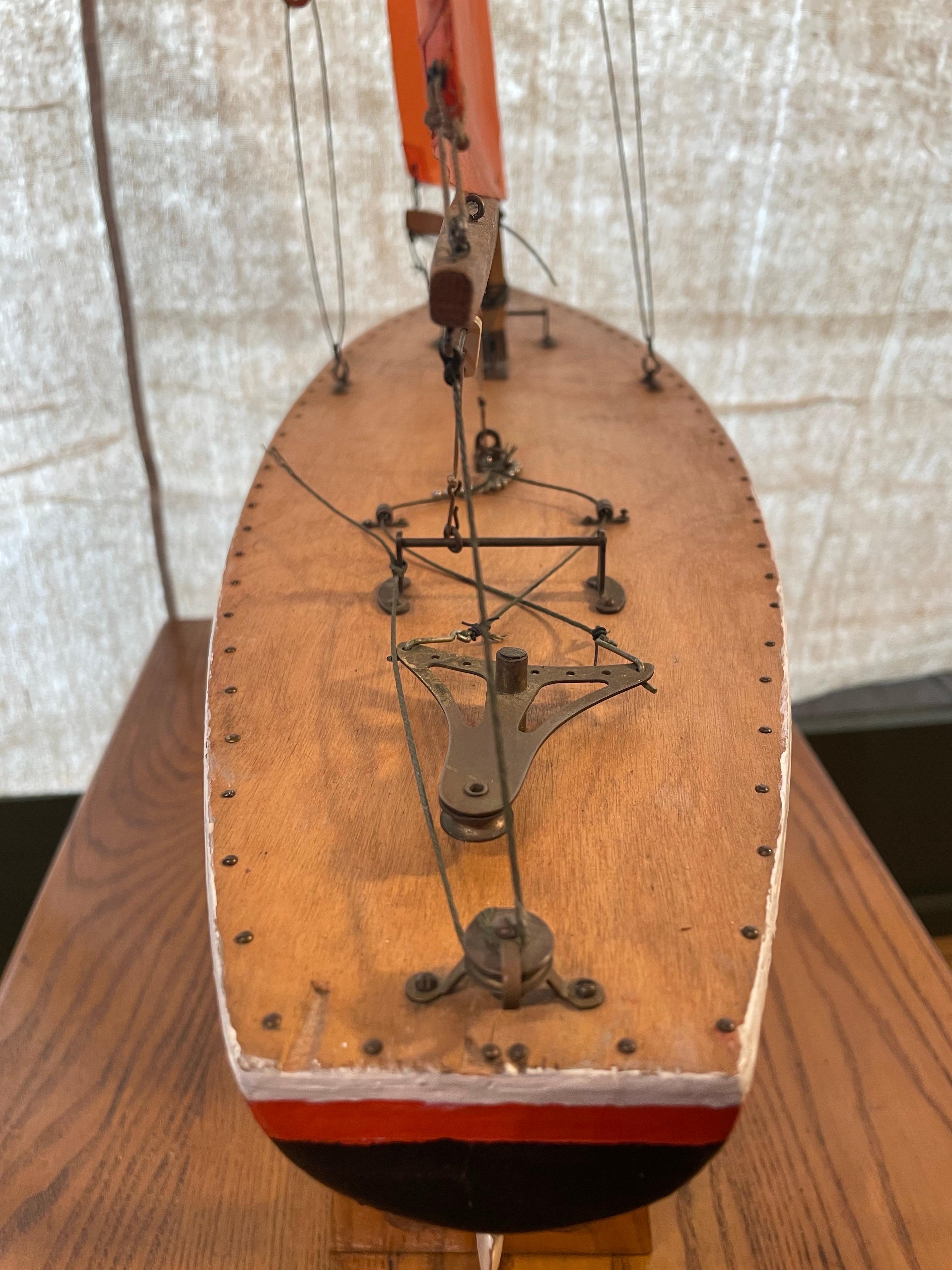 Un charmant yacht artisanal, datant probablement du milieu du 20e siècle, doté d'une coque en bois aux lignes de flottaison élégantes et peintes en rouge et blanc. Le modèle présente un élégant gréement de sloop avec des voiles rouges aux couleurs