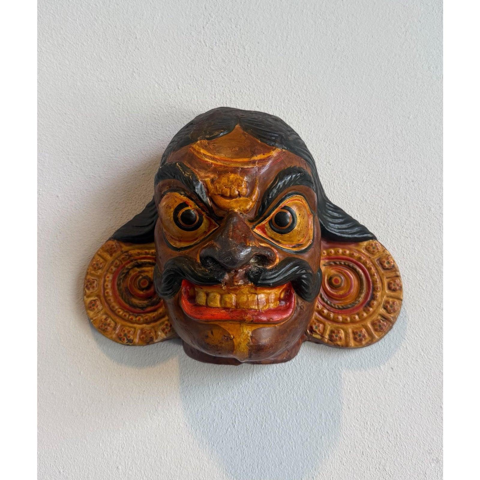 Questa maschera dipinta è un pezzo sorprendente ed espressivo, scolpito in modo audace e dai colori vivaci, probabilmente ispirato a disegni teatrali o rituali tradizionali del Sud o del Sud-Est asiatico. Il volto, fuso in una base ocra profonda,