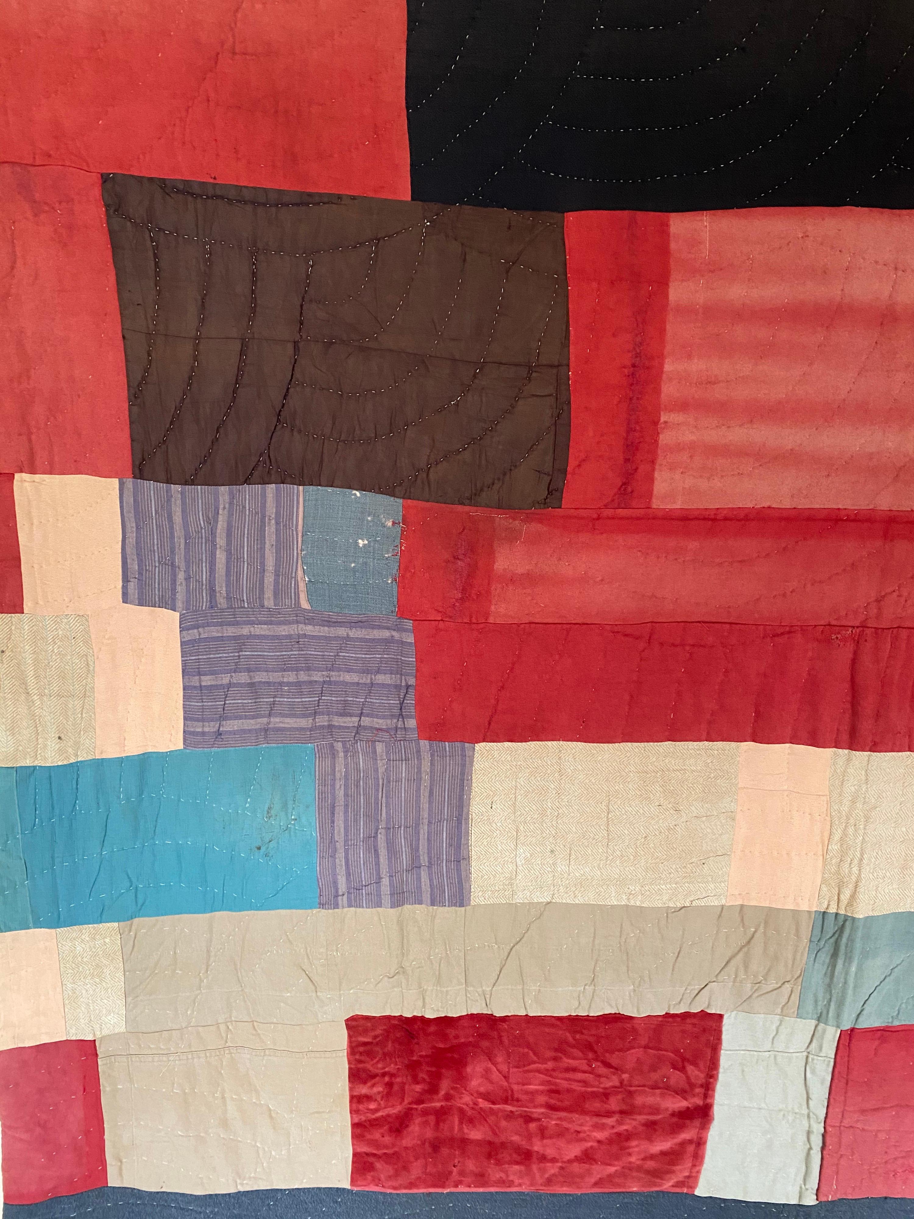 A vintage American early 20th century patchwork quilt. Consisting of primarily red block-work with contrasting shades of blue, cream, and dark grays. Cream colored quilted backing.