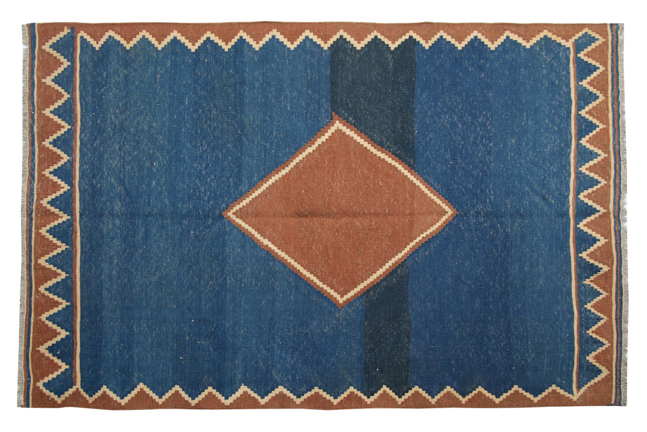 This elegant handwoven wool area rug features a simple color palette of rust, cream and blue. The central medallion is a simple diamond woven in rust and is surrounded by a beautiful open blue field and a zig-zag cream and rust border. Both the