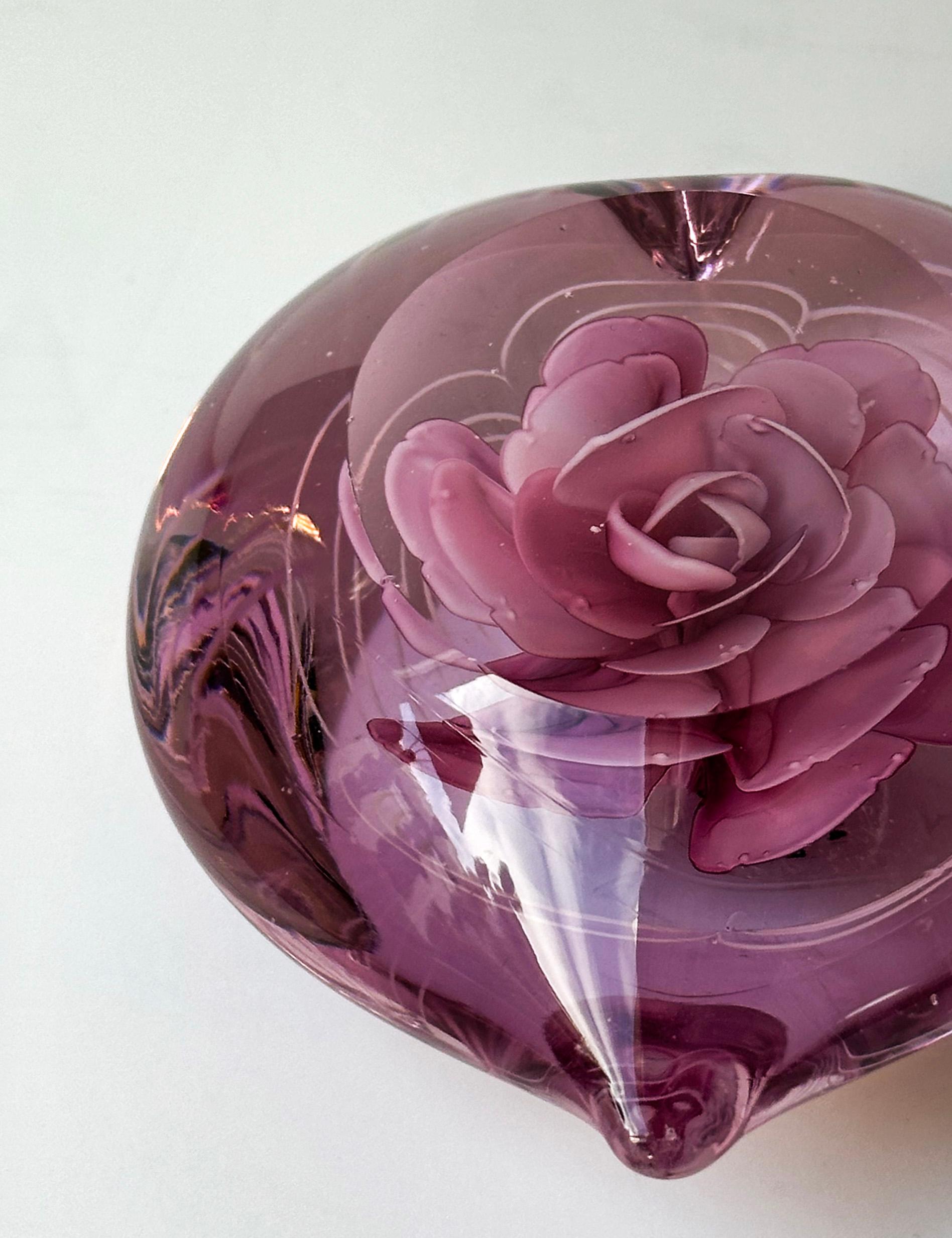 This vintage pink glass paperweight, meticulously crafted and shaped in the form of a love heart, radiates charm with its brilliantly bright pink glass. Encased within are delicate glass rose petals, framed by finely looped lines, showcasing an