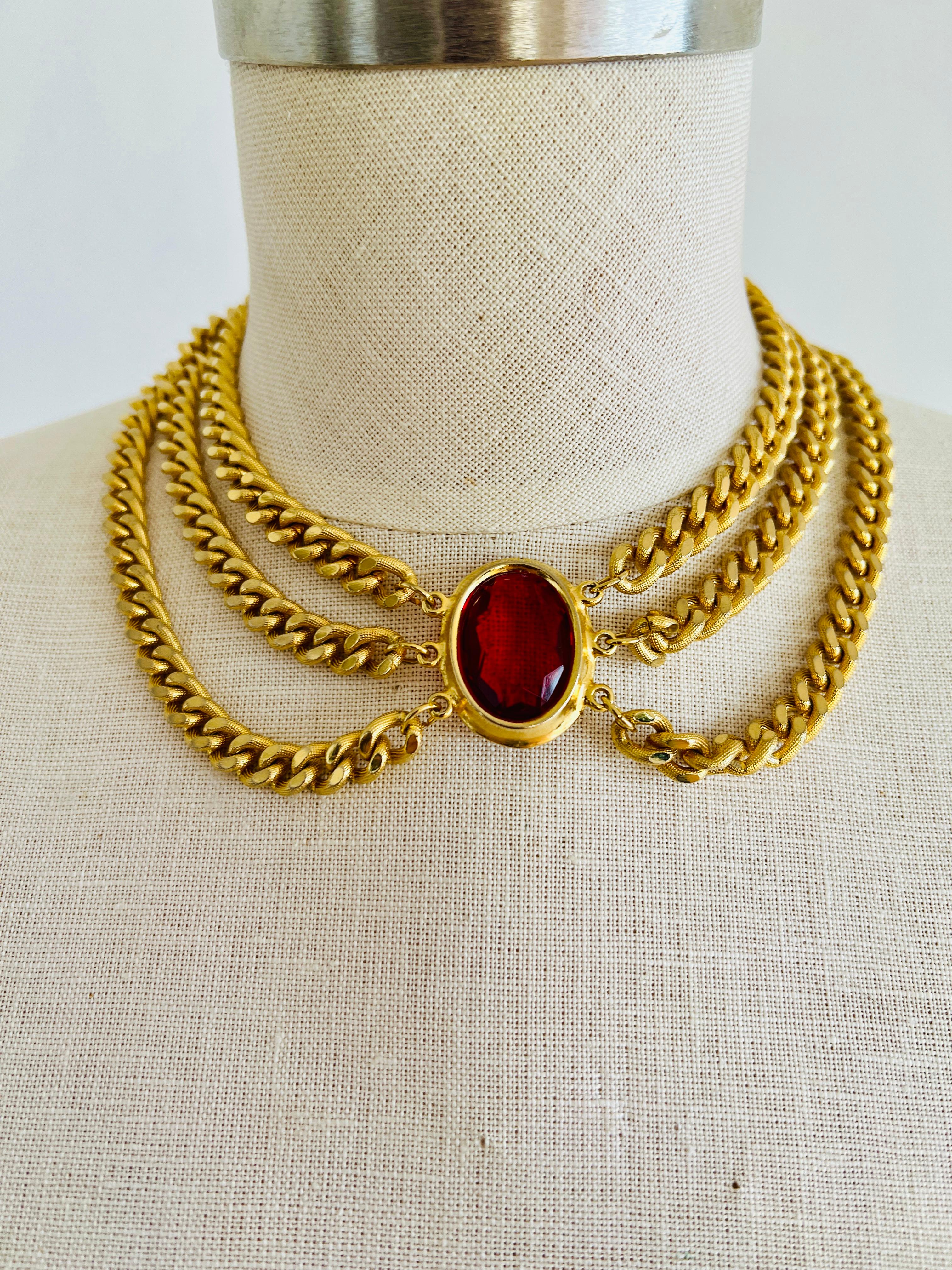 Discover the allure of this runway-inspired choker statement necklace, a composition of three weighty, gold-plated Cuban chains, converging at a striking faceted red glass oval jewel centerpiece. This necklace promises to be the highlight of any