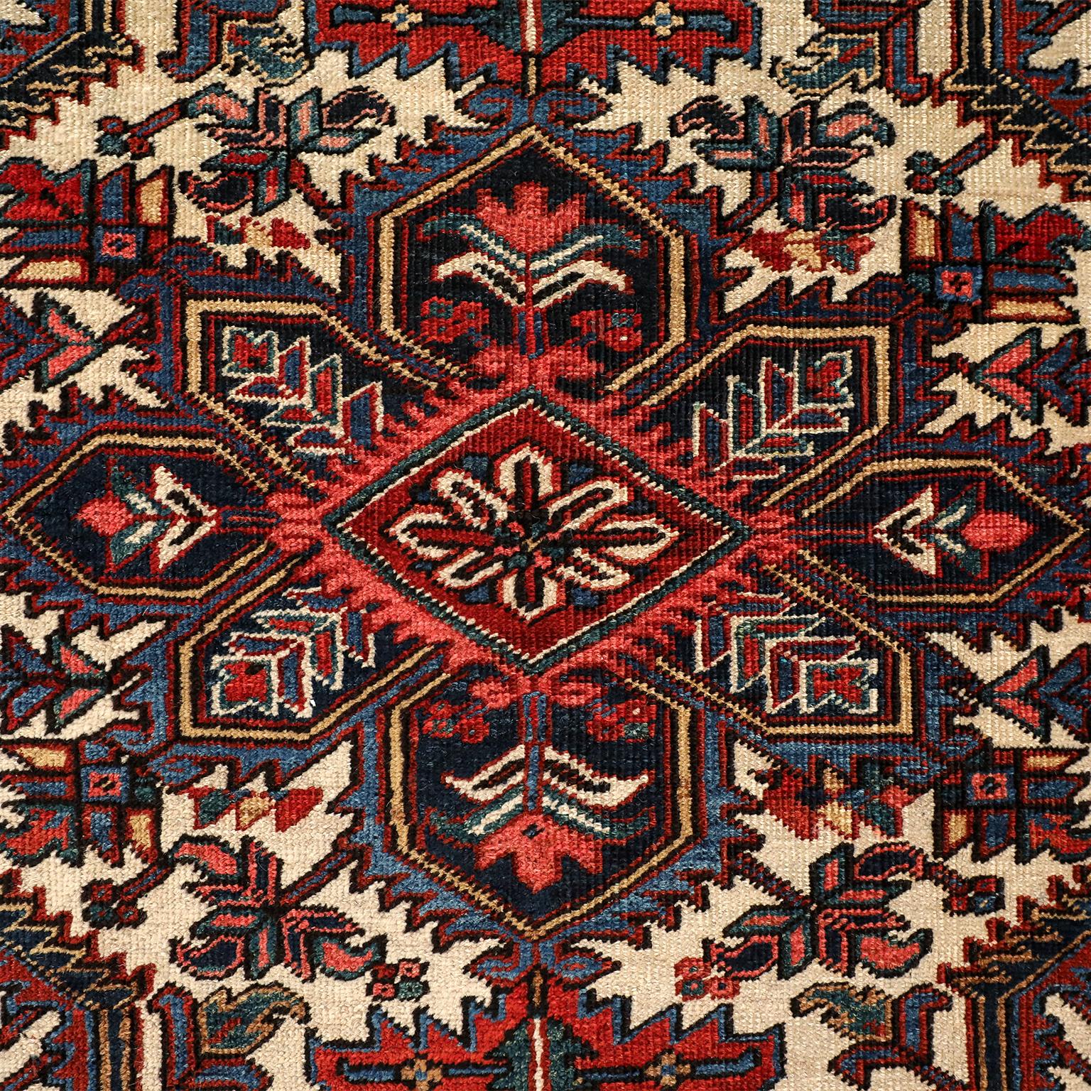 This vintage Heriz Persian carpet circa 1920s in pure wool and vegetable dyes is hand knotted using a Persian knot upon a cotton warp and weft. It features a brightly colored geometric floral medallion upon a cream-colored field, with a