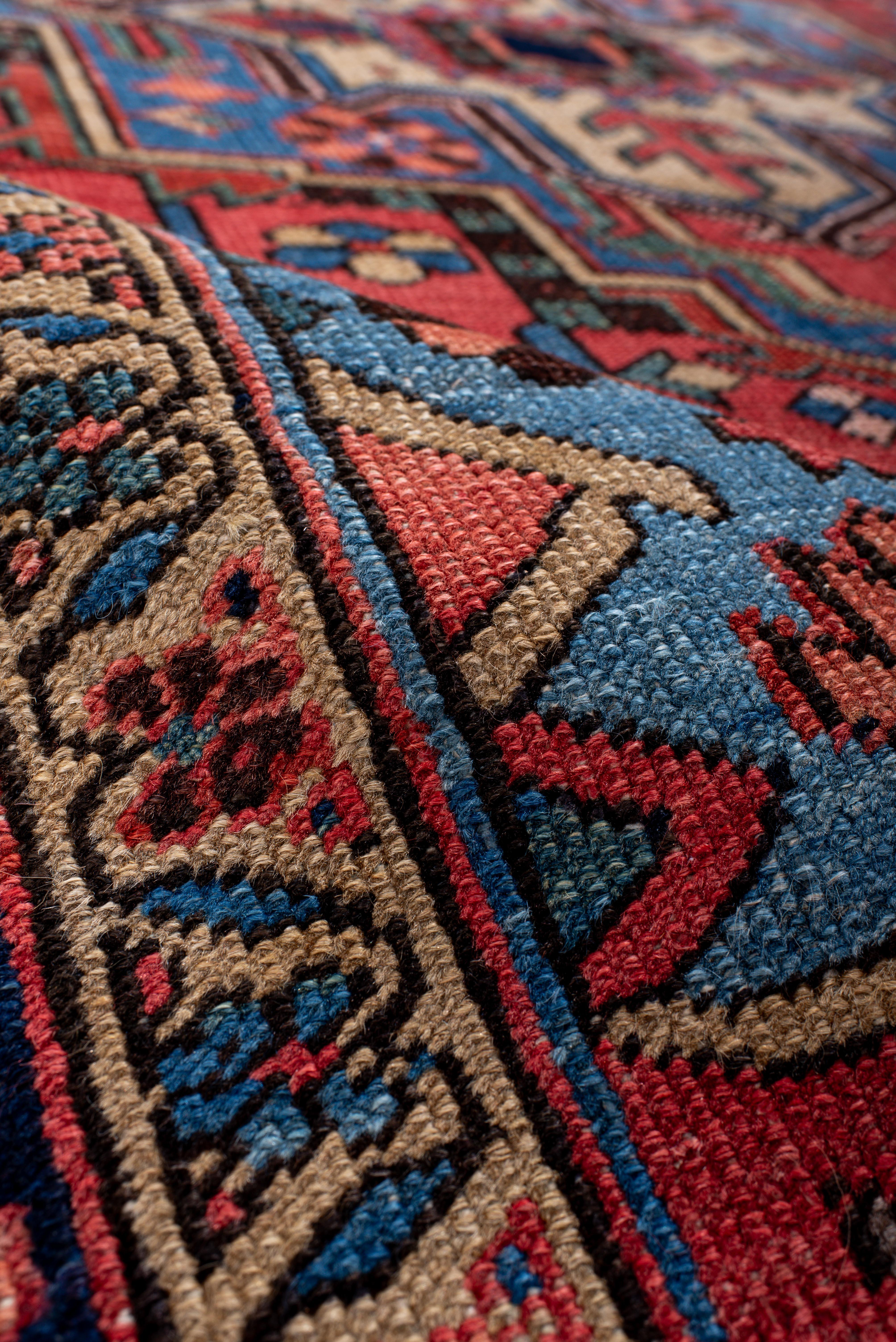 Dieser Vintage-Teppich von Heriz strotzt vor Energie und zeigt ein sattes, karminrotes Feld, das von einem kühnen, kantigen Medaillon in Himmelblau, Elfenbein und Schwarz dominiert wird. Die Komposition ist mit stilisierten Blumenköpfen,