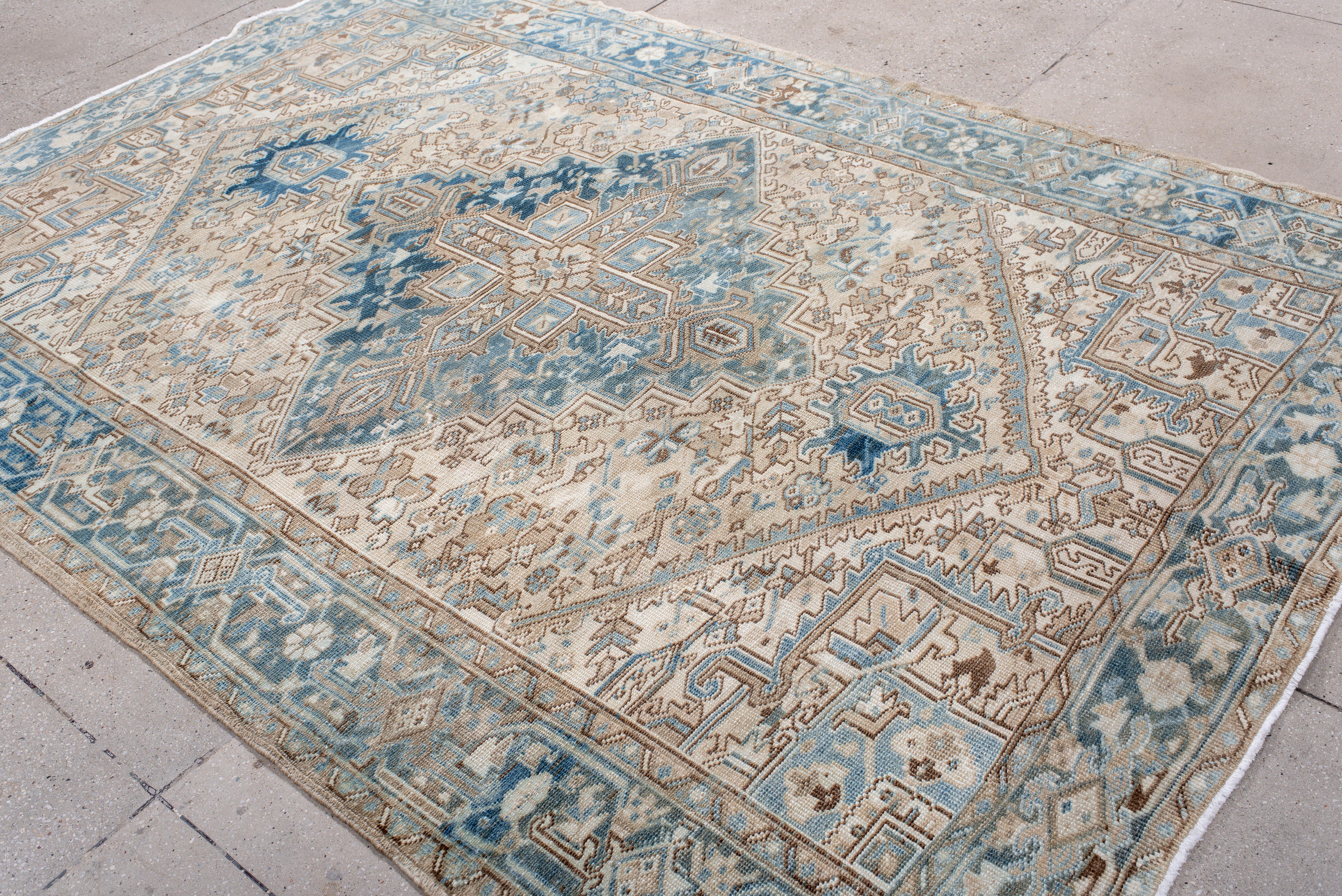 Esta alfombra Heriz vintage muestra un llamativo medallón central en capas de tonos azul vaquero, cielo y mineral, sobre un fondo crema suavemente desteñido. El campo está lleno de abstracciones florales angulosas y hojas dentadas, formando una