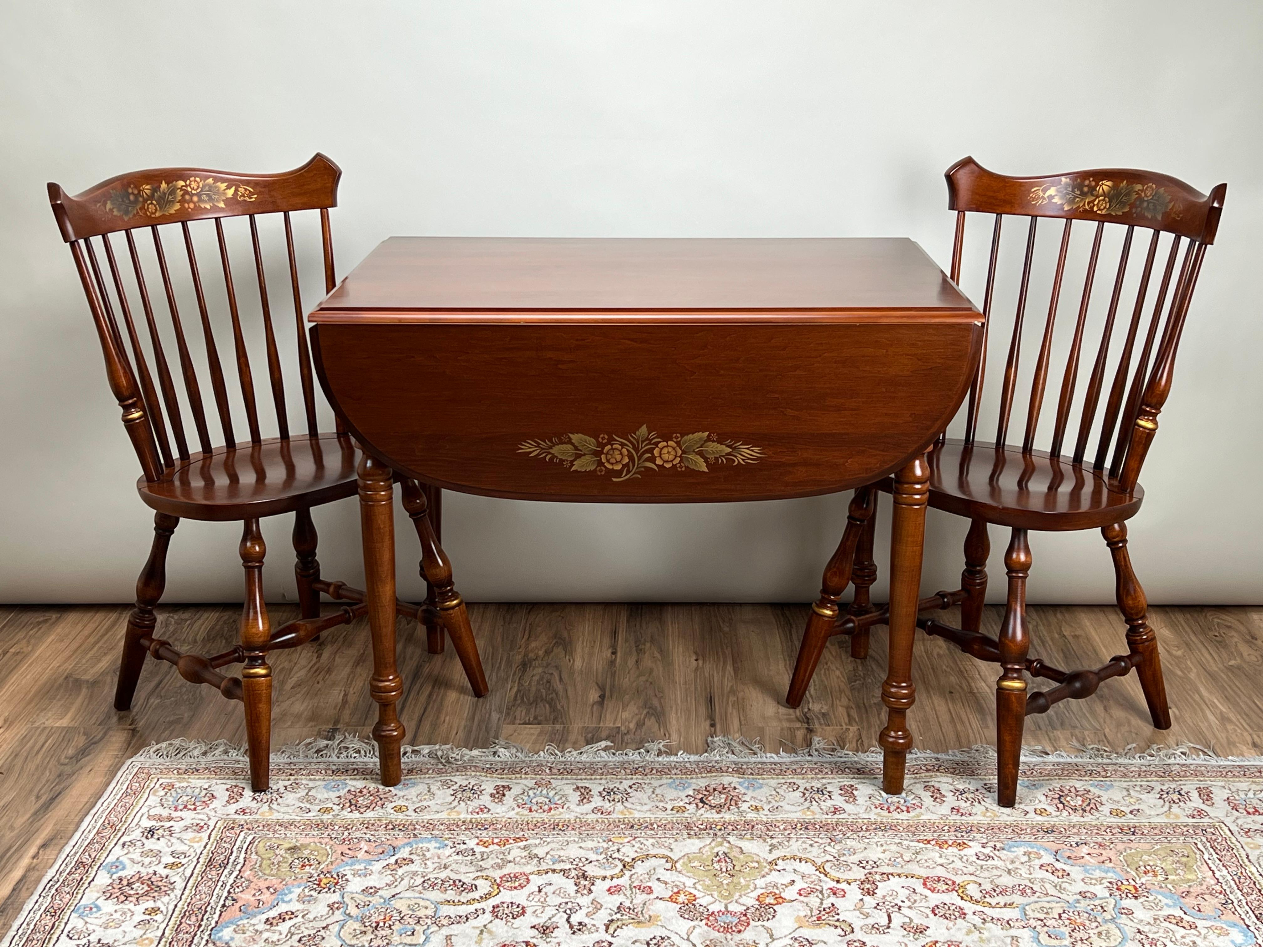 This charming vintage Hitchcock maple breakfast table with matching side chairs offers a perfect blend of traditional New England craftsmanship and timeless appeal. The drop-leaf table features beautiful hand-painted floral detailing on both the