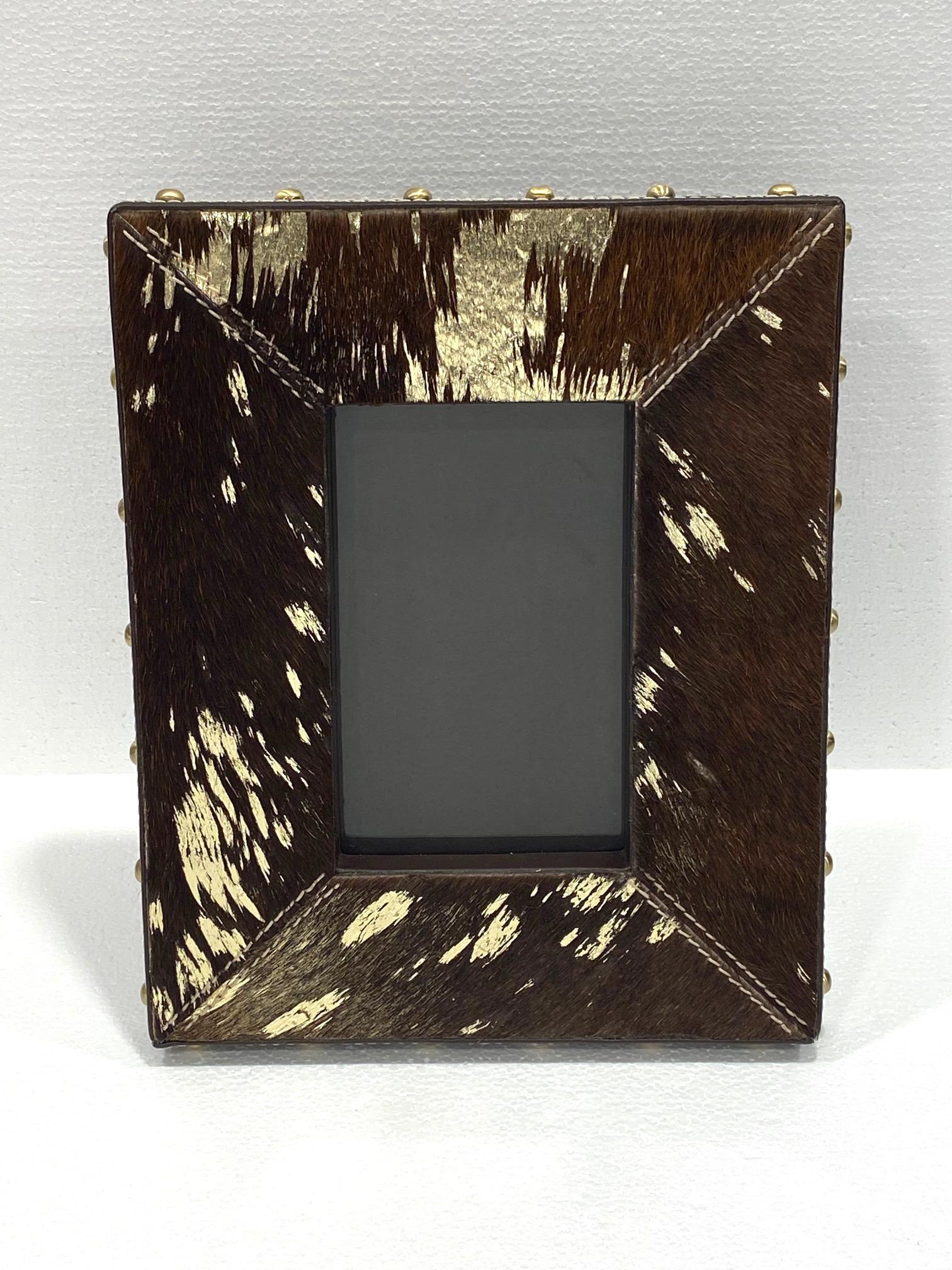 Rustic Hollywood Regency photo frame in dark brown cowhide with gold metallic foil accents. Handcrafted with brown leather trim and brass studs along the borders. Frame features handstitched details and holds 4