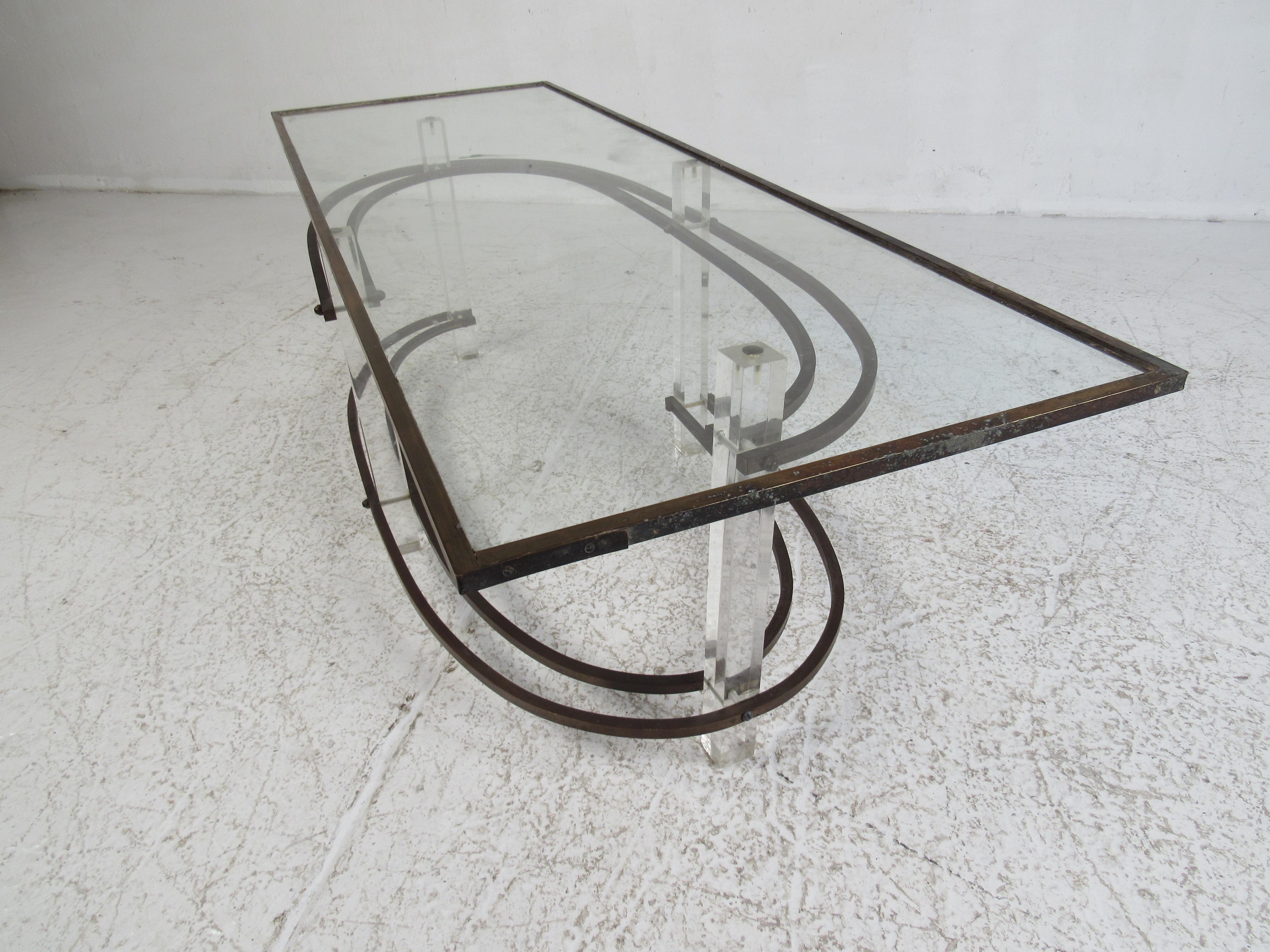 An elegant Mid-Century Modern coffee table designed in the style of Charles Hollis Jones. A unique base with swirling brass supports that stagger and connect to the cubed Lucite legs. A rectangular glass top within a brass and acrylic frame. This