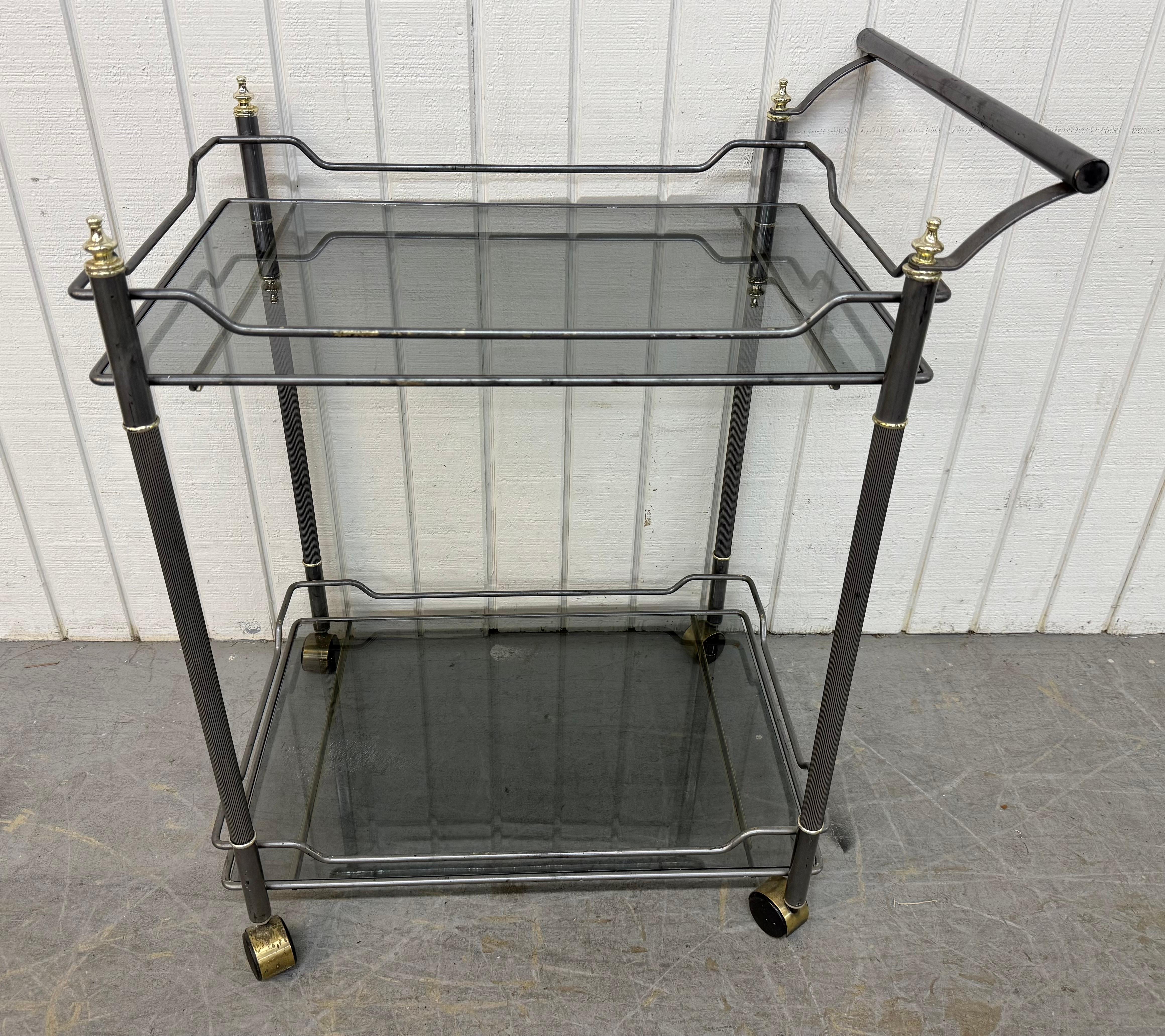 Cette annonce concerne un chariot de service Vintage Hollywood Regency. Elle est dotée d'un cadre chromé, de deux étagères en verre fumé pour le rangement, d'une poignée pour faciliter le transport, d'embouts en laiton, de roulettes d'origine et