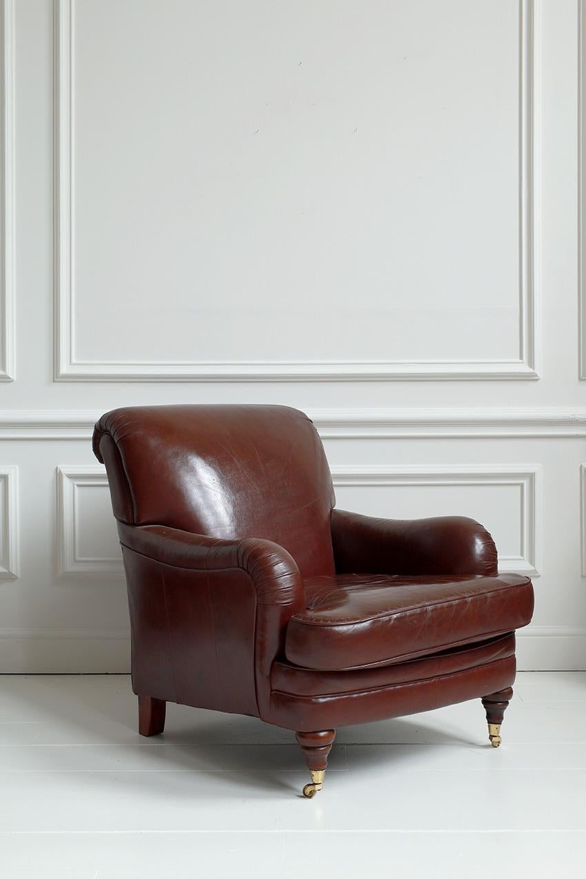 A vintage Howard-style armchair, characterised by its deep seat, low profile, and signature rolled arms. Upholstered in rich, brown full-grain leather, the chair features visible patina and natural creases, which enhance its vintage character and