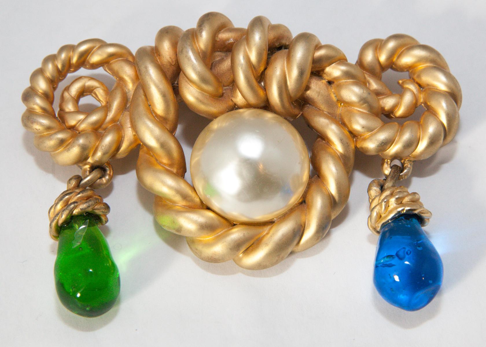 This is a huge, outstanding Chanel brooch.  It has a large twisted rope design with a huge faux pearl center.  Dangling from each side is a large green and blue Gripoix glass drop.  It is in a gold tone setting and measures 4-1/2” wide x 3” long