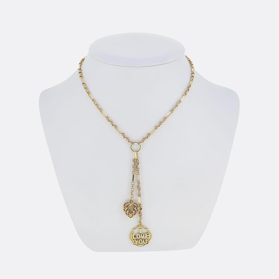 This is a vintage 9ct yellow gold fancy long and short linked necklace. The necklace showcases a pair of matching charms including a filigree styled open love heart and a round charm crafted to read the words 'I Love You'. This necklace is attached