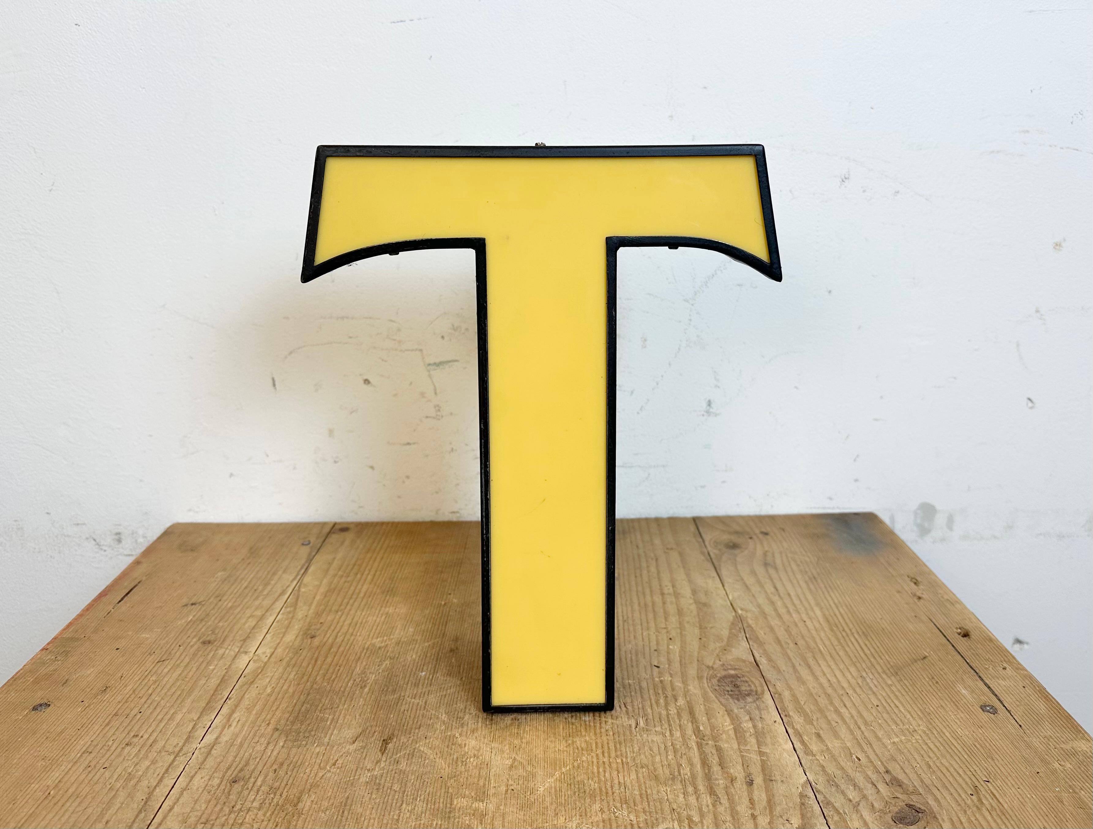 Esta letra T iluminada industrial vintage se fabricó en Italia en la década de 1970 y procede de un antiguo cartel publicitario. Presenta un cuerpo de metal negro y una cubierta de plexiglás amarillo. Está equipado con una tira de LED. La carta