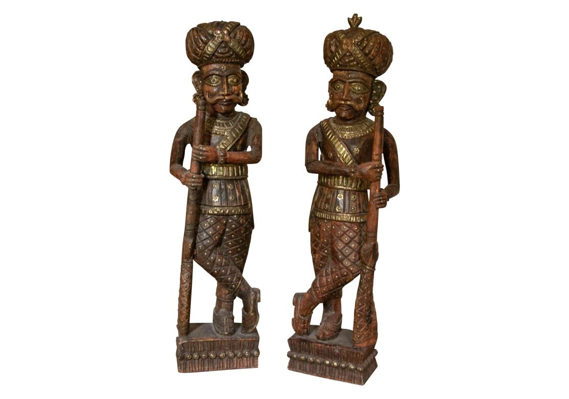 Vintage India Chowkidar wooden statues, hand carved and gilt.  Night watchmen / guards holding on old rifle and staff. The guards are wearing beautifully carved, brass nailhead and gilt decorated attire and a feathered turban.
Dimensions: 31 1/2