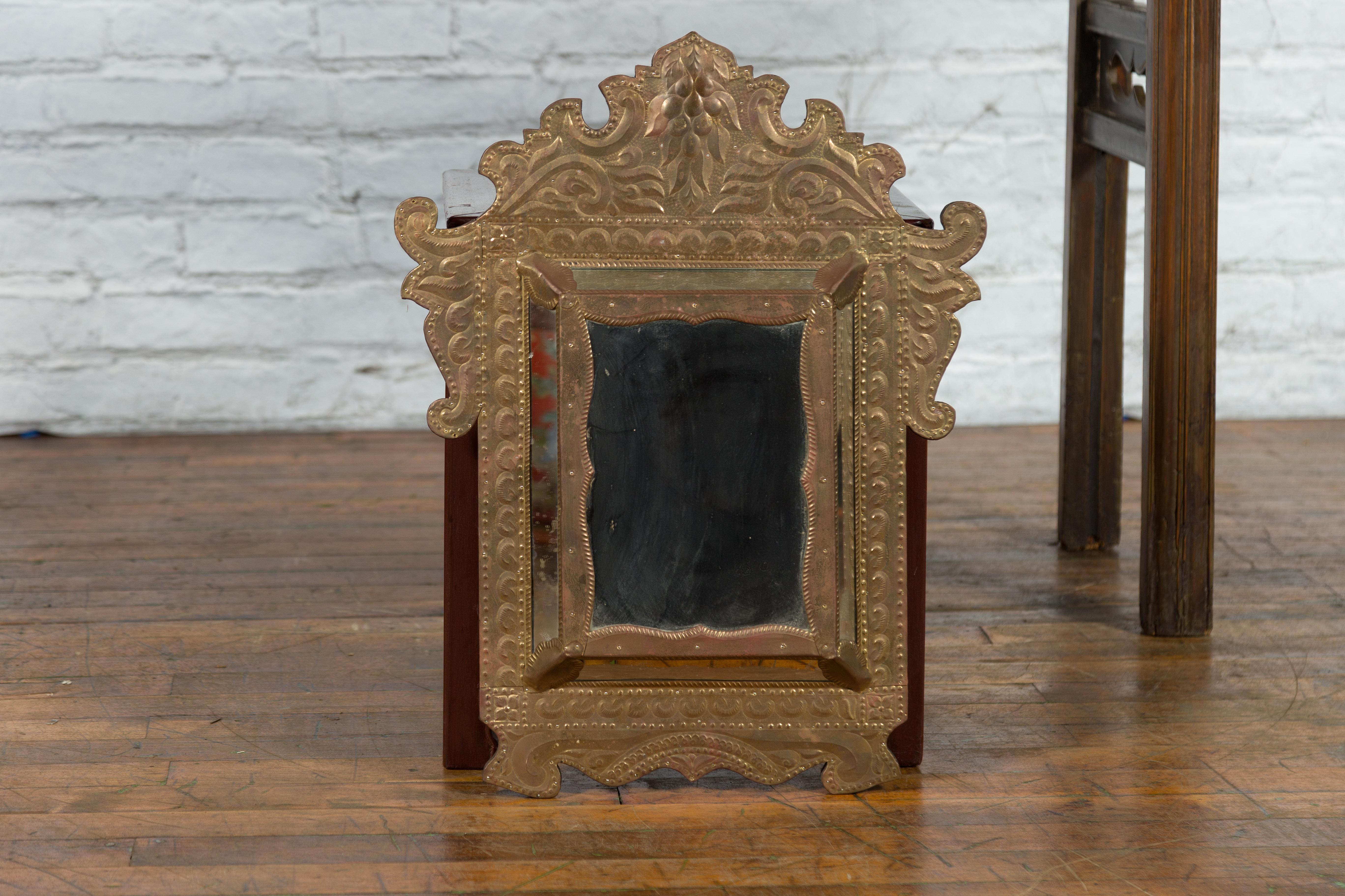 A vintage Indian pareclose mirror from the mid 20th century with gilded metal sheathing over wooden frame. Created in India during the Midcentury period, this small mirror attracts our attention with its ornate crest and golden colors. Showcasing a