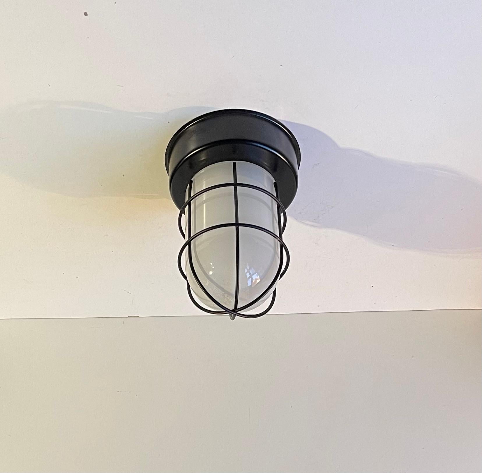A cage flush mount ceiling or wall light in opaline glass and black painted metal. Great industrial or even submarine vibe to it. Suitable for rooms with a low ceiling hight, bathrooms, studios etc. It was designed and made by Abo Randers in Denmark