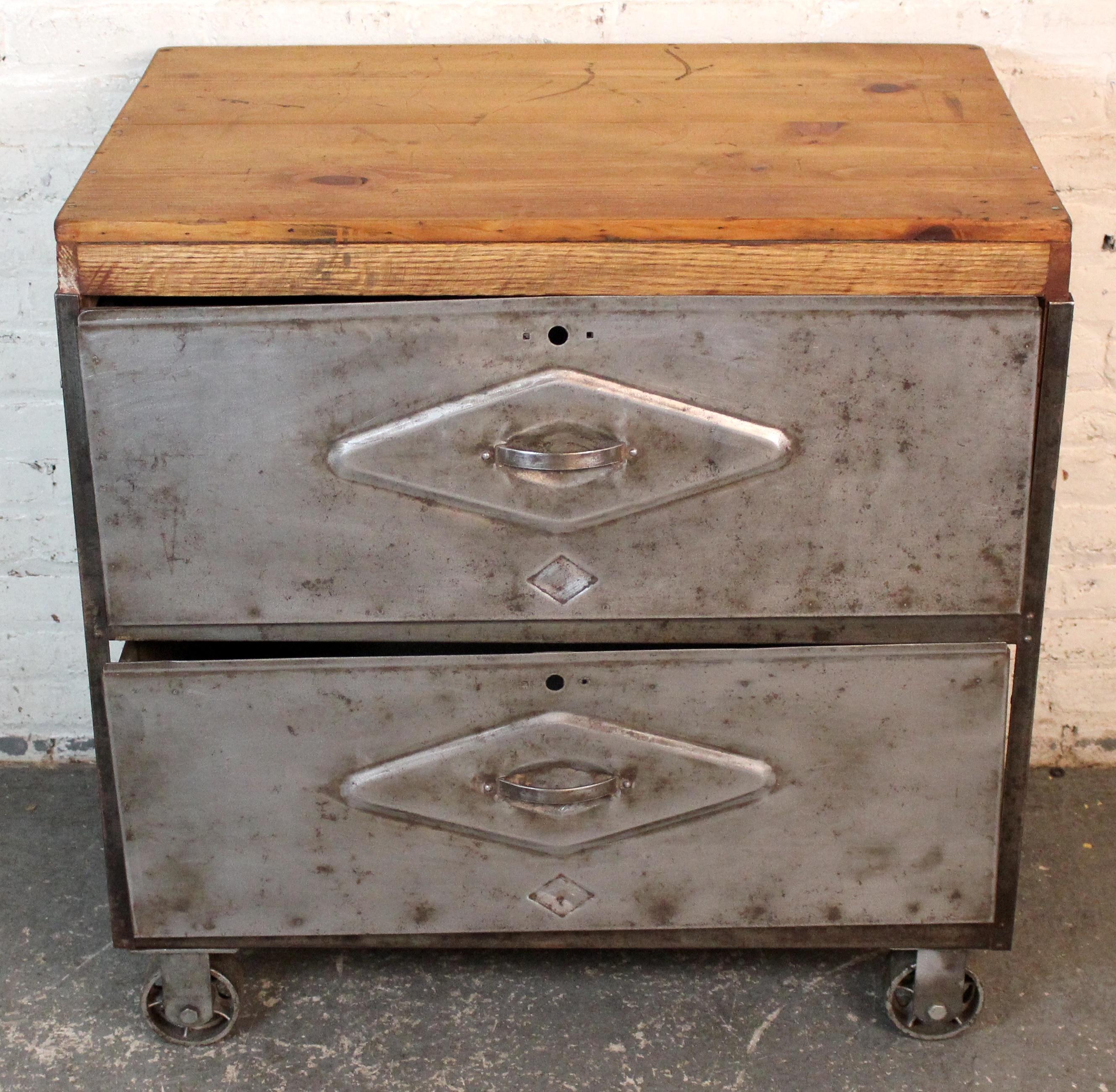 A rugged industrial cart featuring a sturdy metal frame with two oversized drawers and a rich, patinated wood top. Mounted on heavy-duty casters for mobility, this utilitarian piece offers both storage and surface functionality. Perfect as a mobile