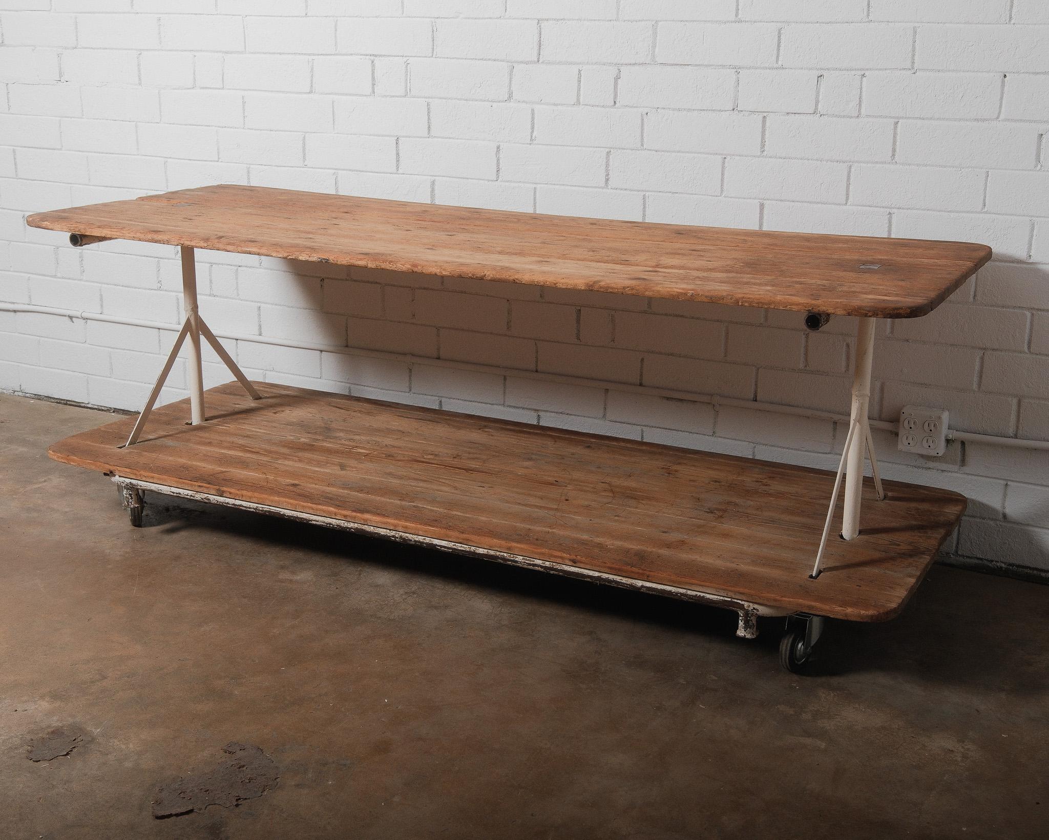 This vintage industrial factory cart table is a striking piece that blends utilitarian design with rustic charm. The solid pine plank top and bottom shelf have a beautifully aged patina, showcasing rich wood grain, natural imperfections, and signs