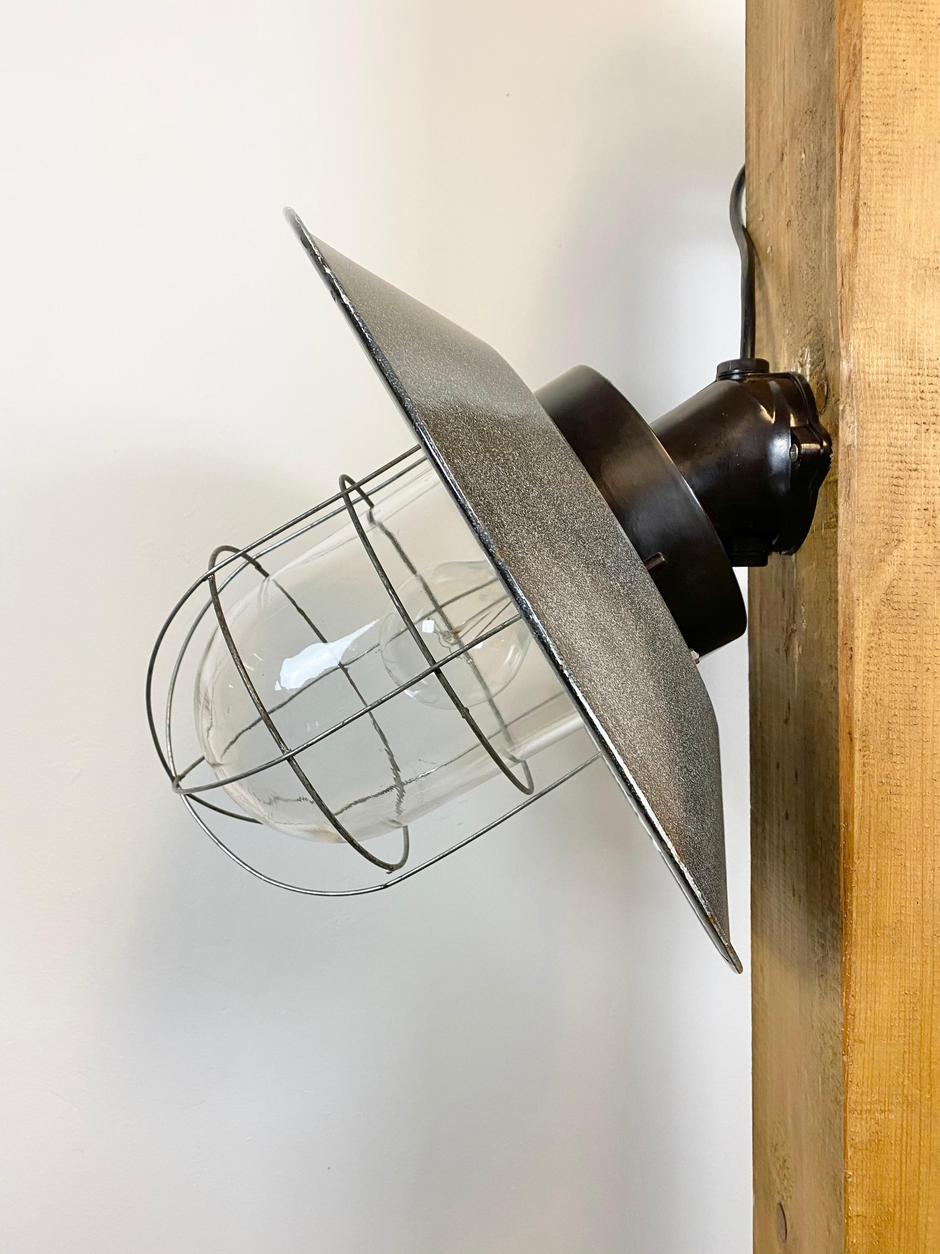- Industrial wall lamp with bakelite wall mounting from former Czechoslovakia
- Made during the 1960s
- Grey enamel shade with white interior
- Clear glass, iron grid
- Porcelain socket for E 27 lightbulbs and new wire
- Weight: 3.5 kg.