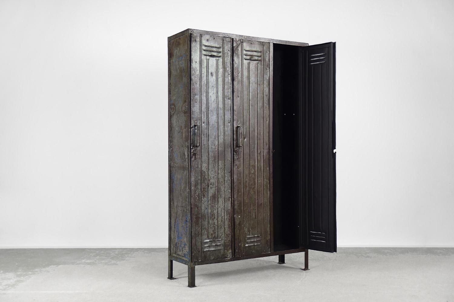 This industrial, metal factory cabinet was produced during the 1950s. The wardrobe has three doors with ventilation openings. There are hangers for clothes inside. Layers of paints and varnishes, combined with natural tarnish, create a