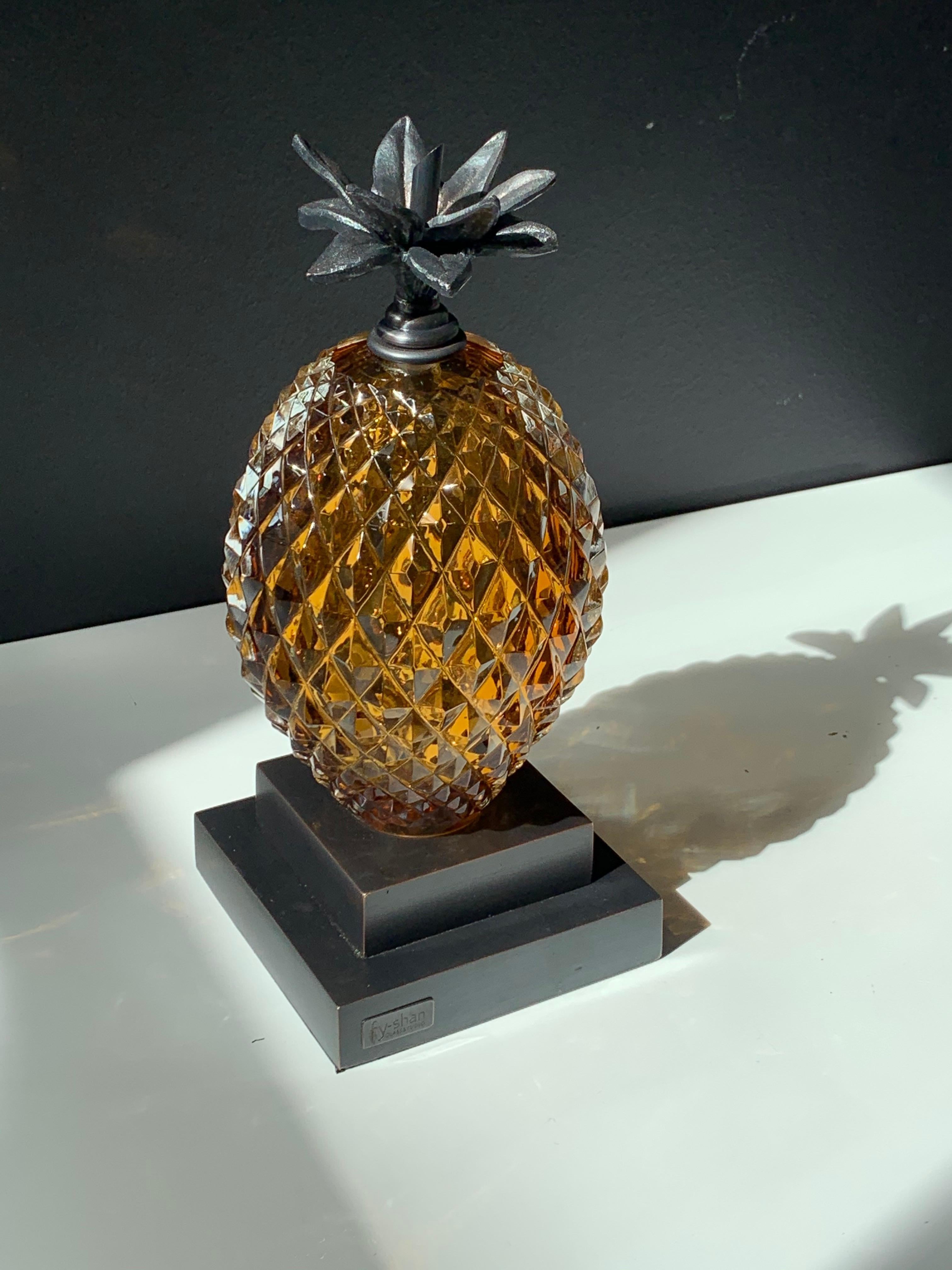 Mundgeblasene und handgeschnitzte Ananas aus bernsteinfarbenem Glas in Verbindung mit einem Ständer aus Gussmetall und einem Blattmotiv, das als Buchstütze oder als Akzent in zeitlosen Innenräumen funktioniert.