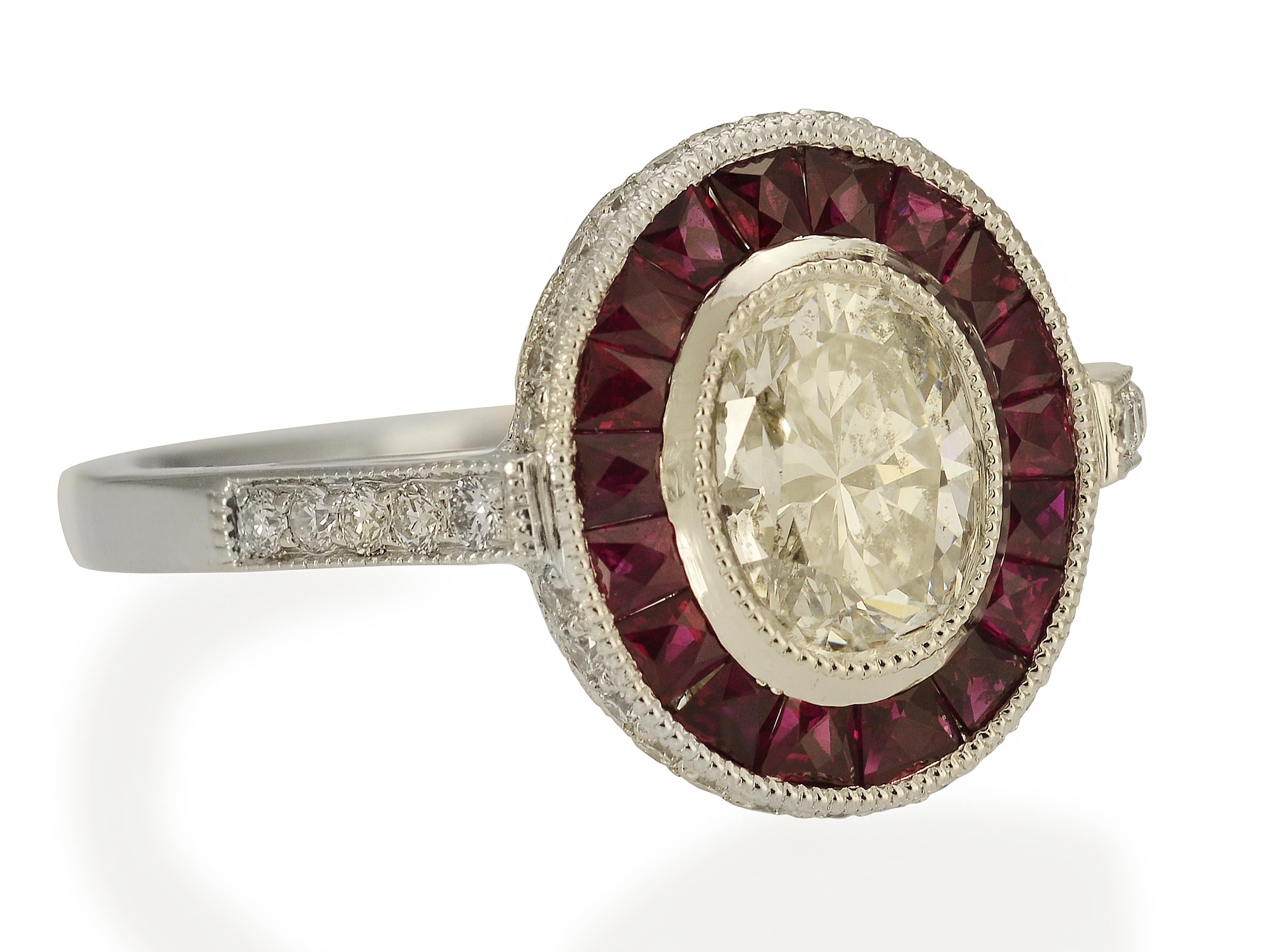This art deco inspired ring is handmade in platinum. It features a bezel-set 0.77 carat Oval diamond of H color and SI2 clarity in the center. Surrounding the diamond are 18 caliber cut rubies totaling 0.53 carats. An additional 0.22 carats of round