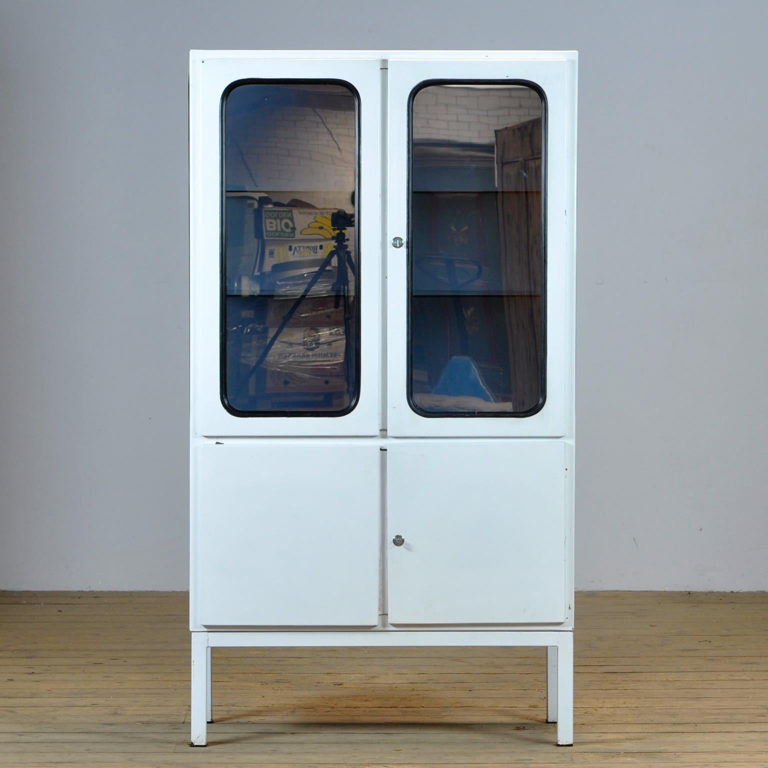 This medical cabinet was designed in the 1970s and was produced circa 1975 in hungary. It is made from iron and glass with new two glass shelves. The glass is semi-transparent/reflective. The glass is held by a black rubber strip. The cabinet