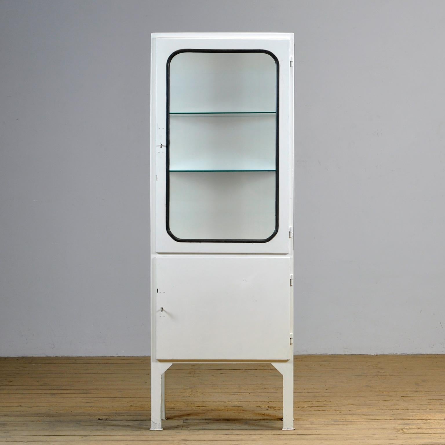 This medical cabinet was designed in the 1970s and was produced circa 1975 in hungary. It is made from iron and glass with new glass shelves. The glass is held by a black rubber strip. The cabinet features two adjustable glass shelves and a