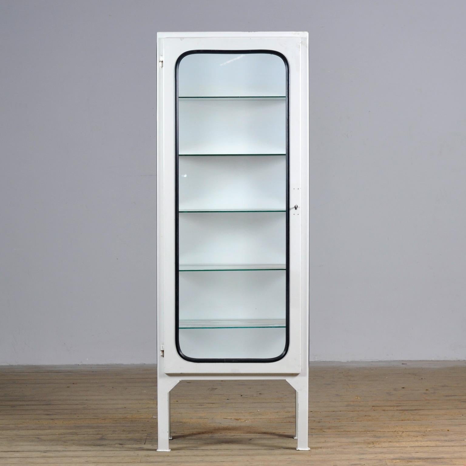 Ce cabinet médical a été conçu dans les années 1970 et a été produit vers 1975 en Hongrie. Elle est faite de fer et de verre avec de nouvelles étagères en verre. Le verre est maintenu par une bande de caoutchouc noir. L'armoire comporte cinq