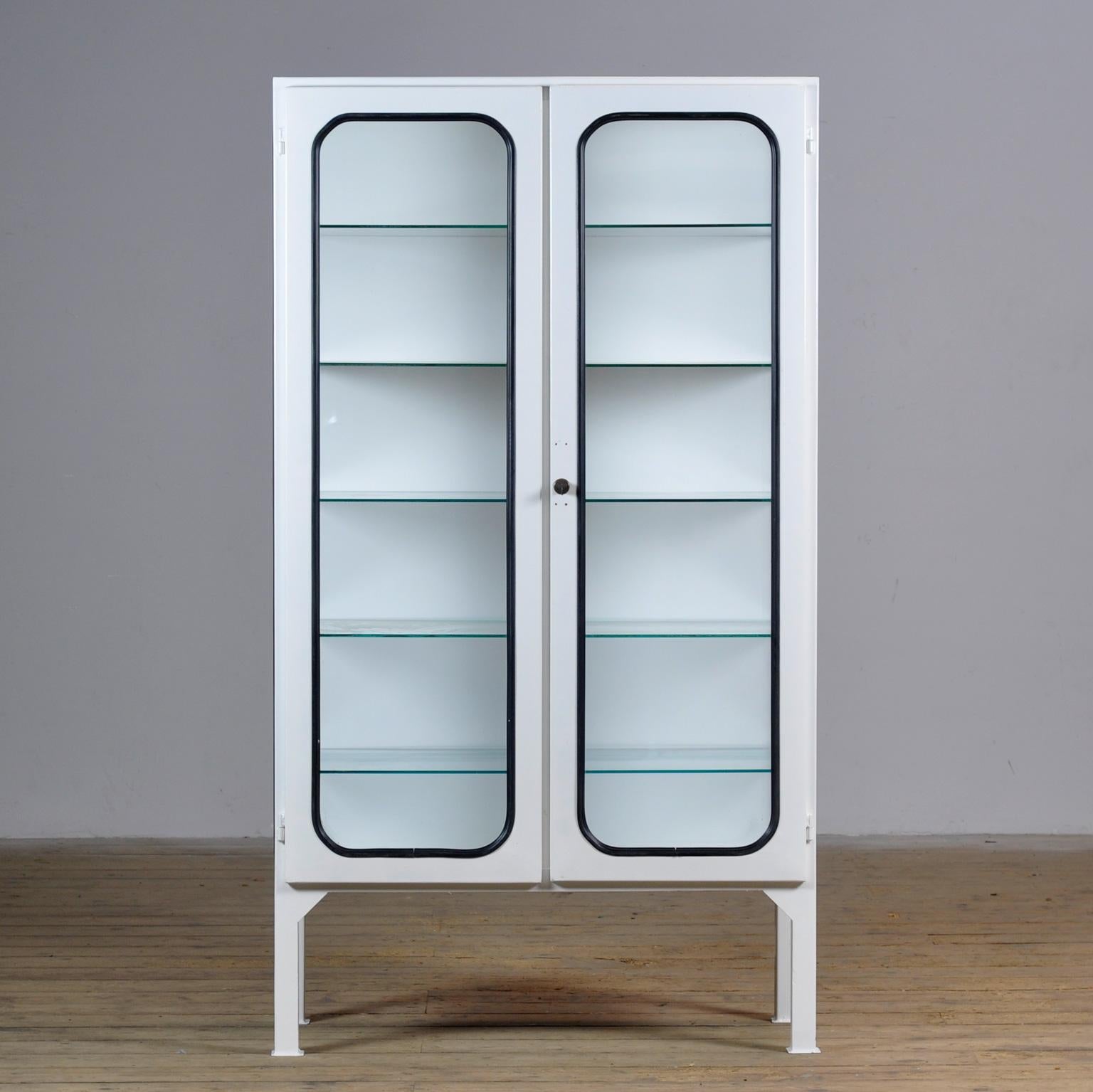Ce cabinet médical a été conçu dans les années 1970 et a été produit vers 1975 en Hongrie. Elle est faite de fer et de verre avec de nouvelles étagères en verre. Le verre est maintenu par une bande de caoutchouc noir. L'armoire comporte cinq