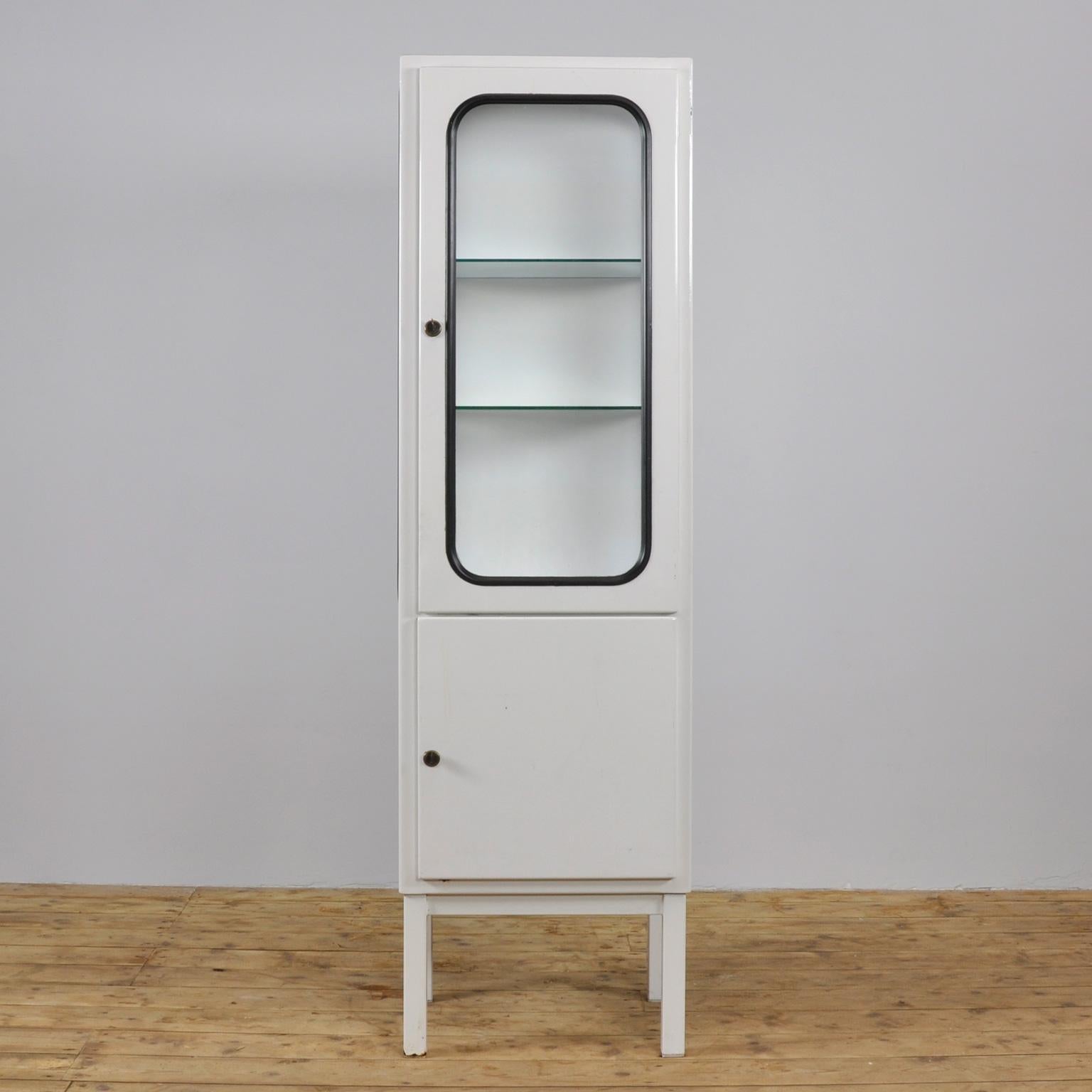 This medical cabinet was designed in the 1970s and produced in 1975, in Hungary. It is made from steel and glass, with the glass panes held in place by a black rubber strip. The cabinet features two adjustable glass shelves and functioning locks.