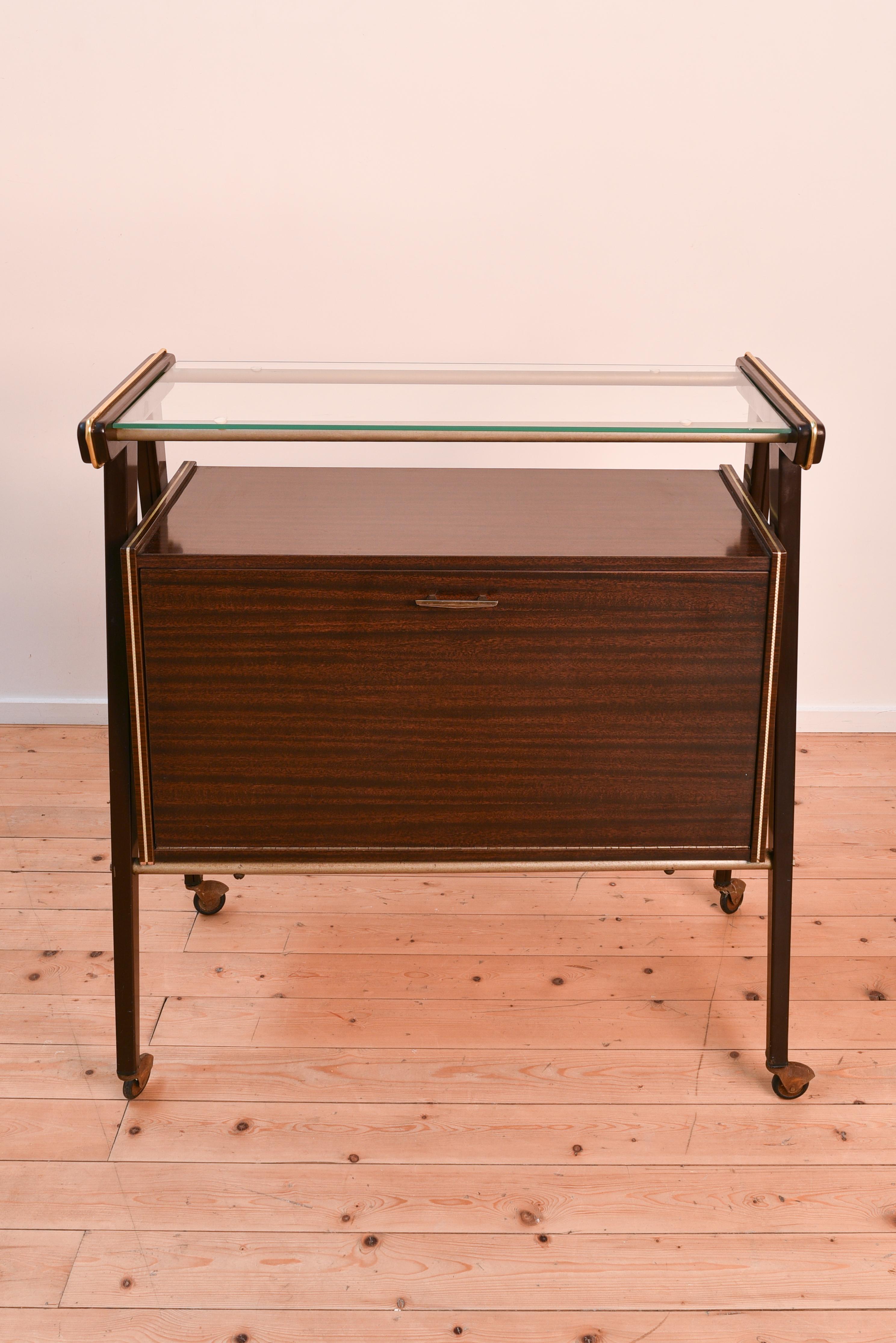 This very elegant bar cart was made in the 1960s by Rama Mobili and has a glass shelf and a cabinet underneath with a little light bulb on the inside. When you open the cabinet door the light automatically switches on. 

The bar cart has some