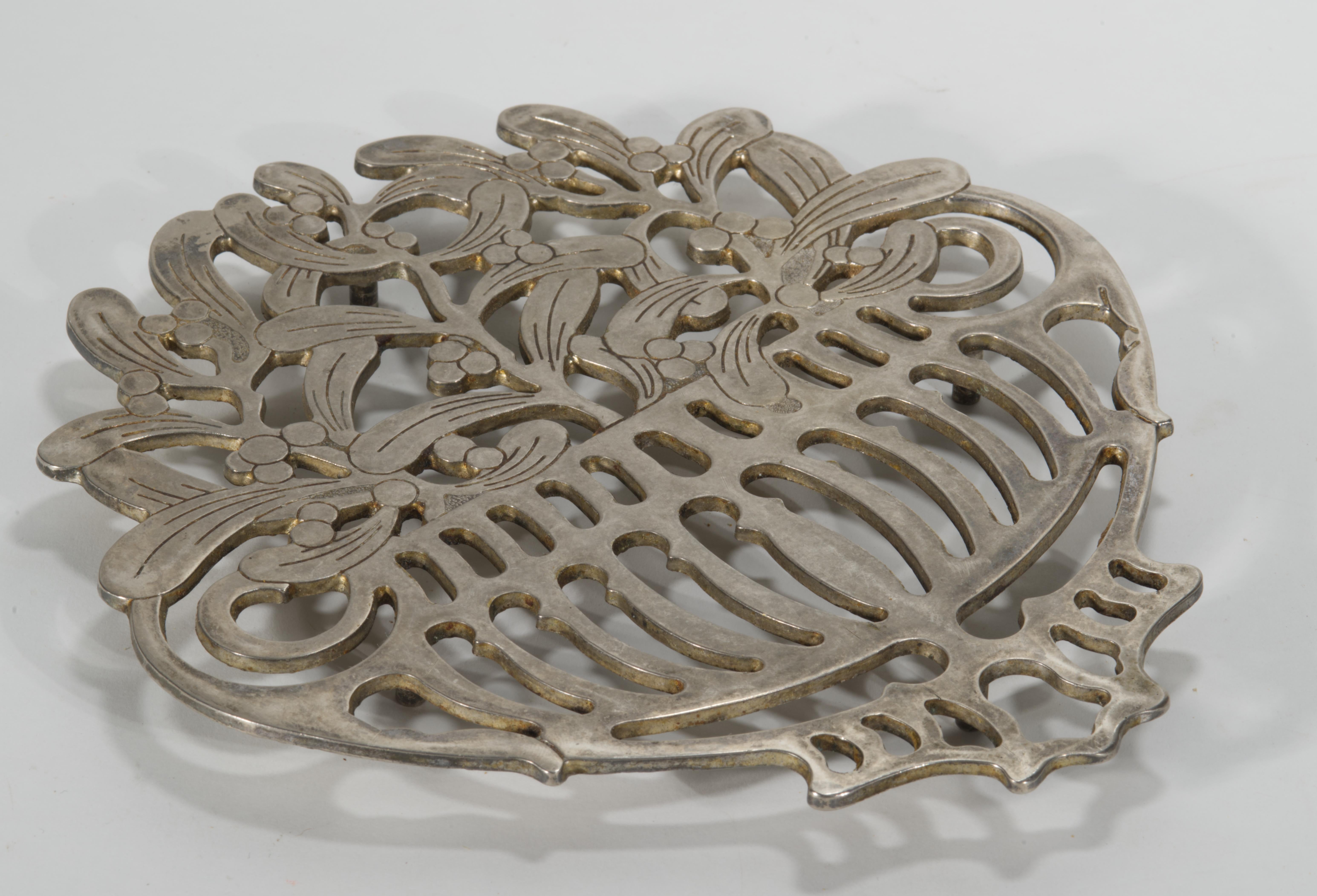 El salvamanteles escultórico de metal presenta un elaborado diseño que representa una cesta ornamentada con asas, llena de ramas; hojas y bayas muy detalladas esculpidas en estilo Art Déco con influencia modernista. Se sostiene sobre 4 pequeños
