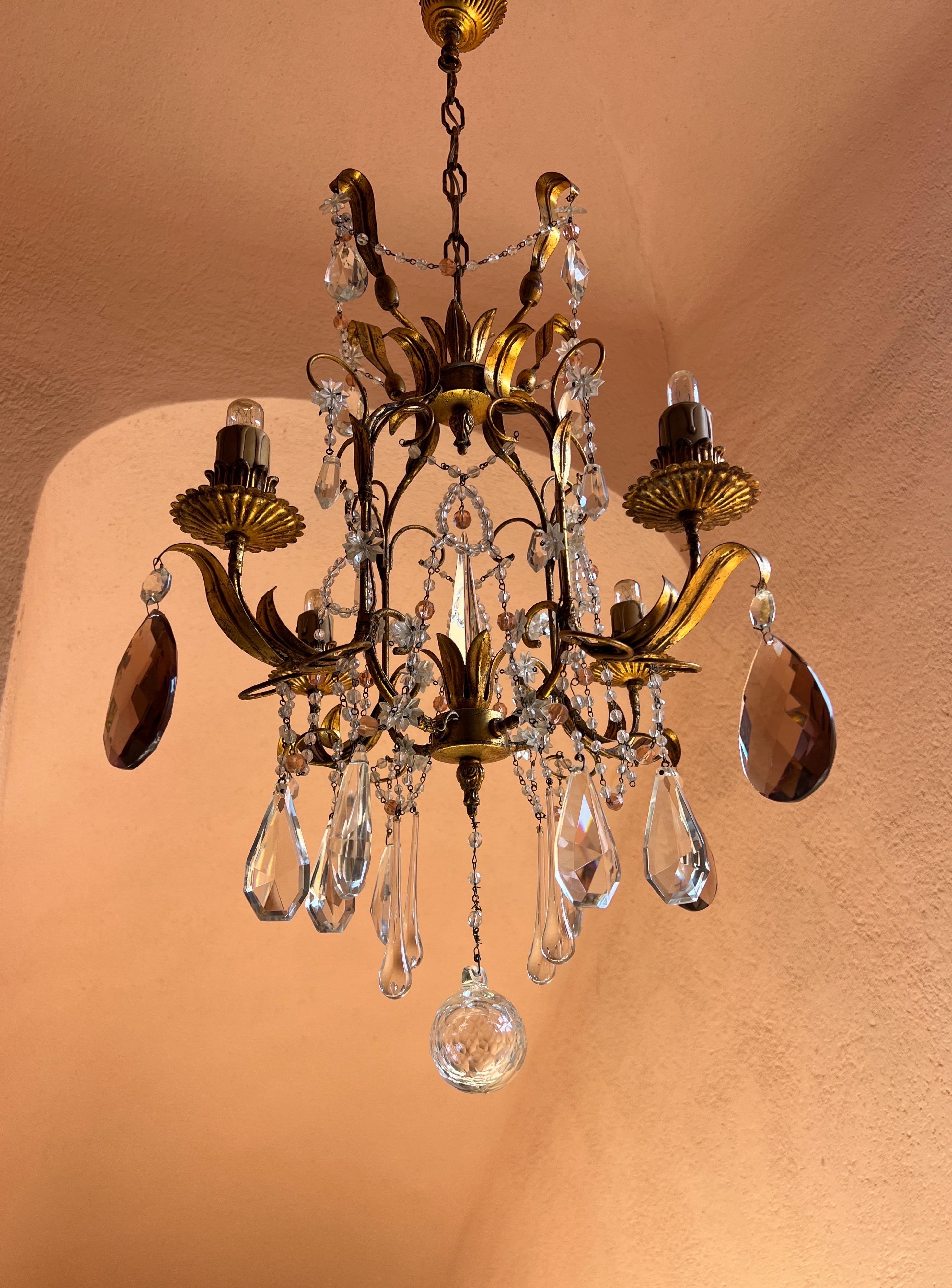 Un elegante lampadario a grappolo a 5 luci ornato da gocce di vetro di Murano nei colori chiaro, rosa e viola. La base in metallo dorato è riccamente decorata con motivi di foglie e dettagli arrotondati che ne esaltano il carattere