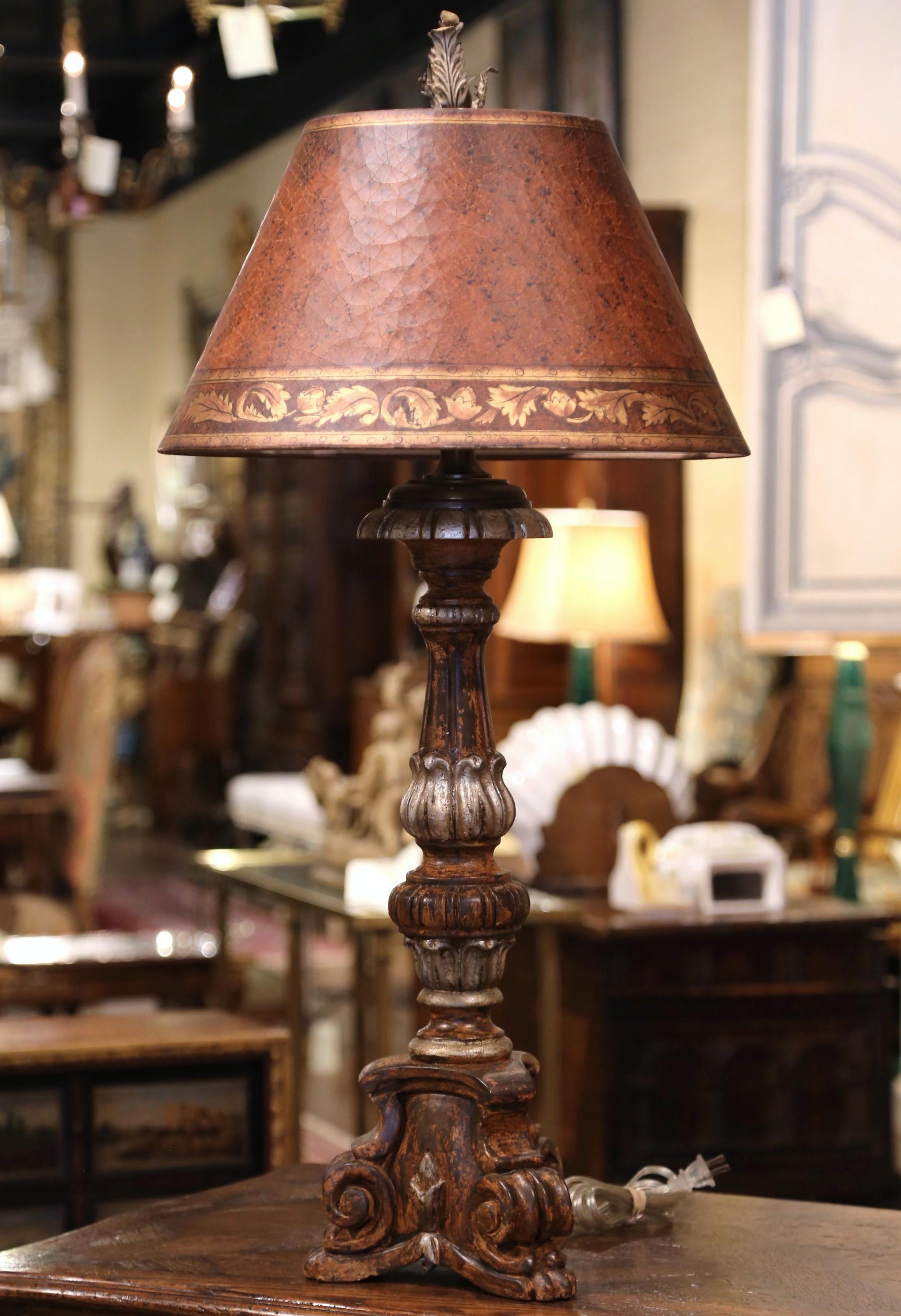 Created in Italy circa 1980, the elegant candlestick stands on scrolled feet over a triangle base decorated with shell decor. The tall carved stem has been fitted into a table lamp with new wiring and is embellished with a brown painted parchment
