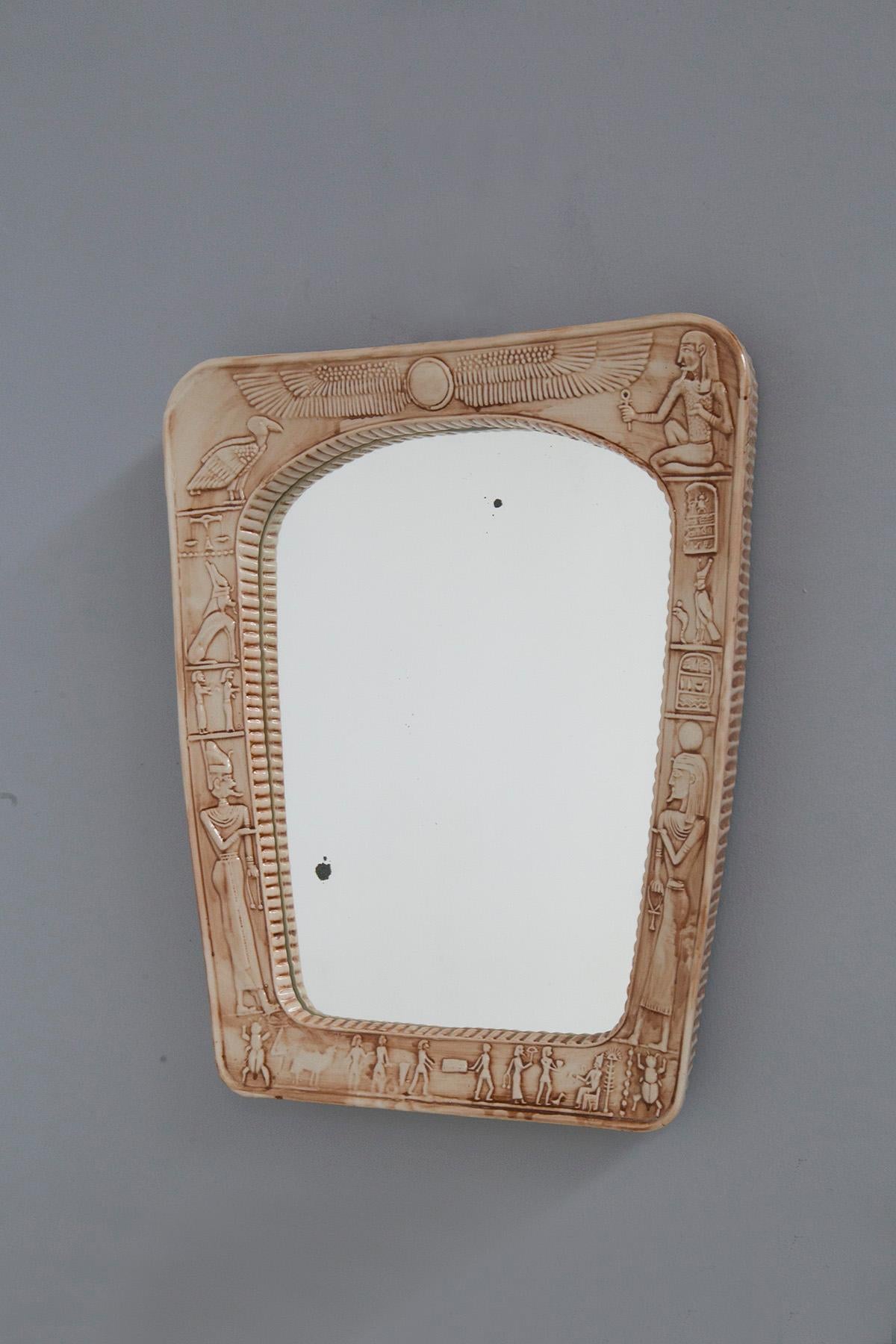 Eccentric vintage Italian mirror made of ceramic. The mirror is circa 1970s. The mirror has a ceramic frame bevelled at the sides, made by hand. Its peculiarity is found on the front, in fact it presents drawings in Egyptian pictographic script. In