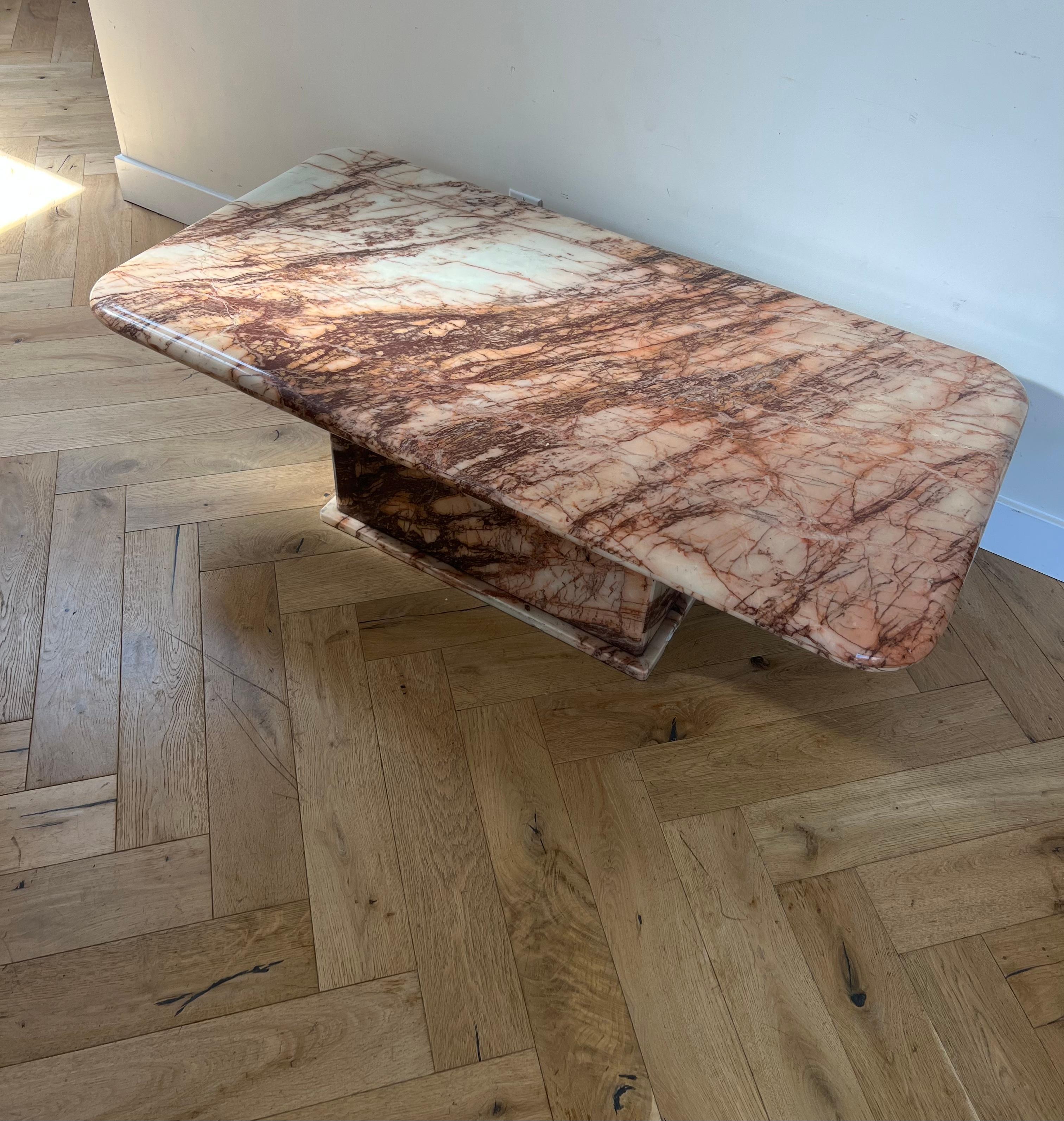 A chunky pink marble coffee table with a very unique burgundy and twig veining, late 1970s. The pedestal base sits atop a small marble platform, while the top features a thick curved edge. Natural signs of age include small moments of scruff to the