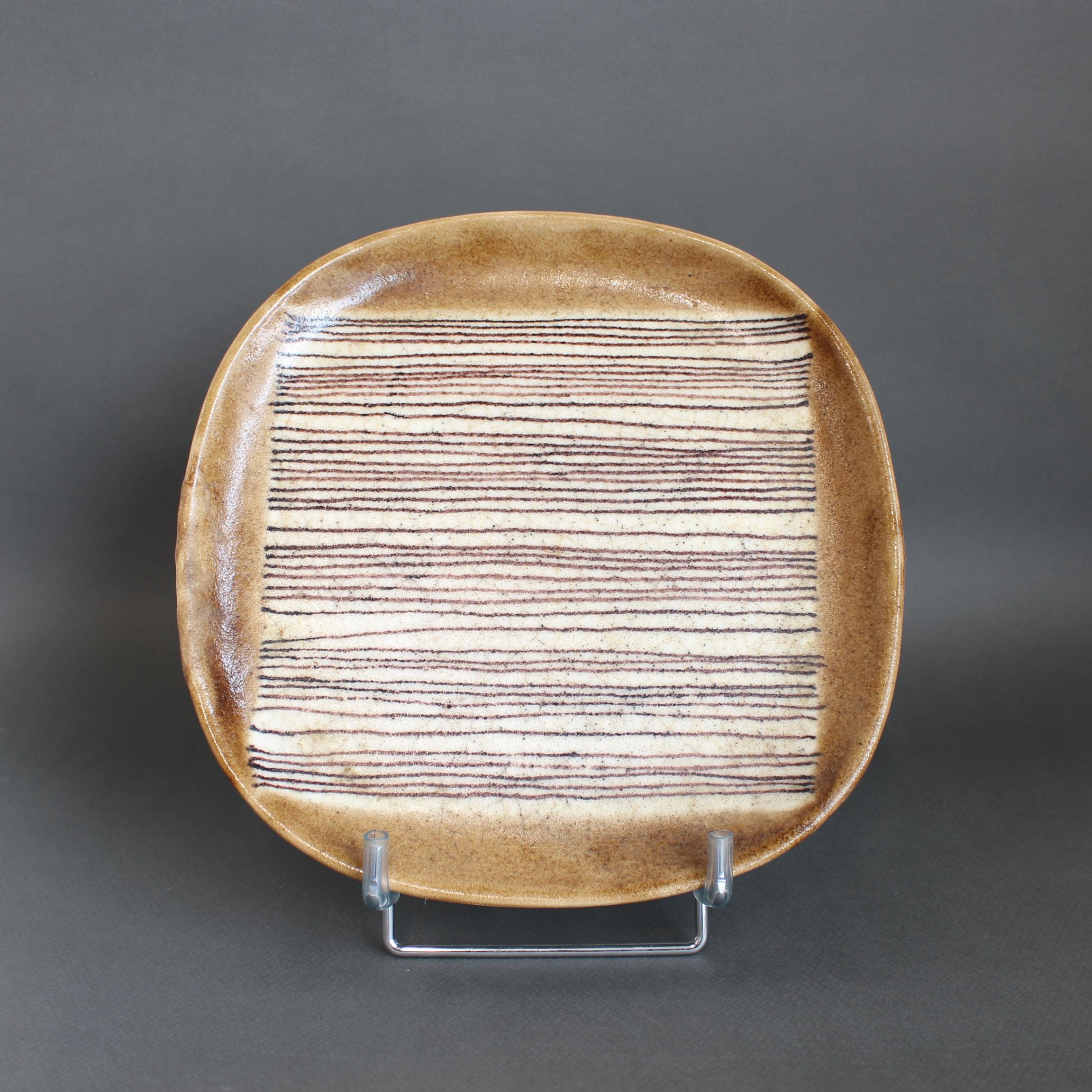 Piatto decorativo in ceramica italiana vintage di Guido Gambone (circa anni '50). Un piatto dai bordi arrotondati dipinto con uno smalto marrone terroso con effetto crepla ai bordi e un motivo a linee orizzontali irregolari al centro. Visivamente,