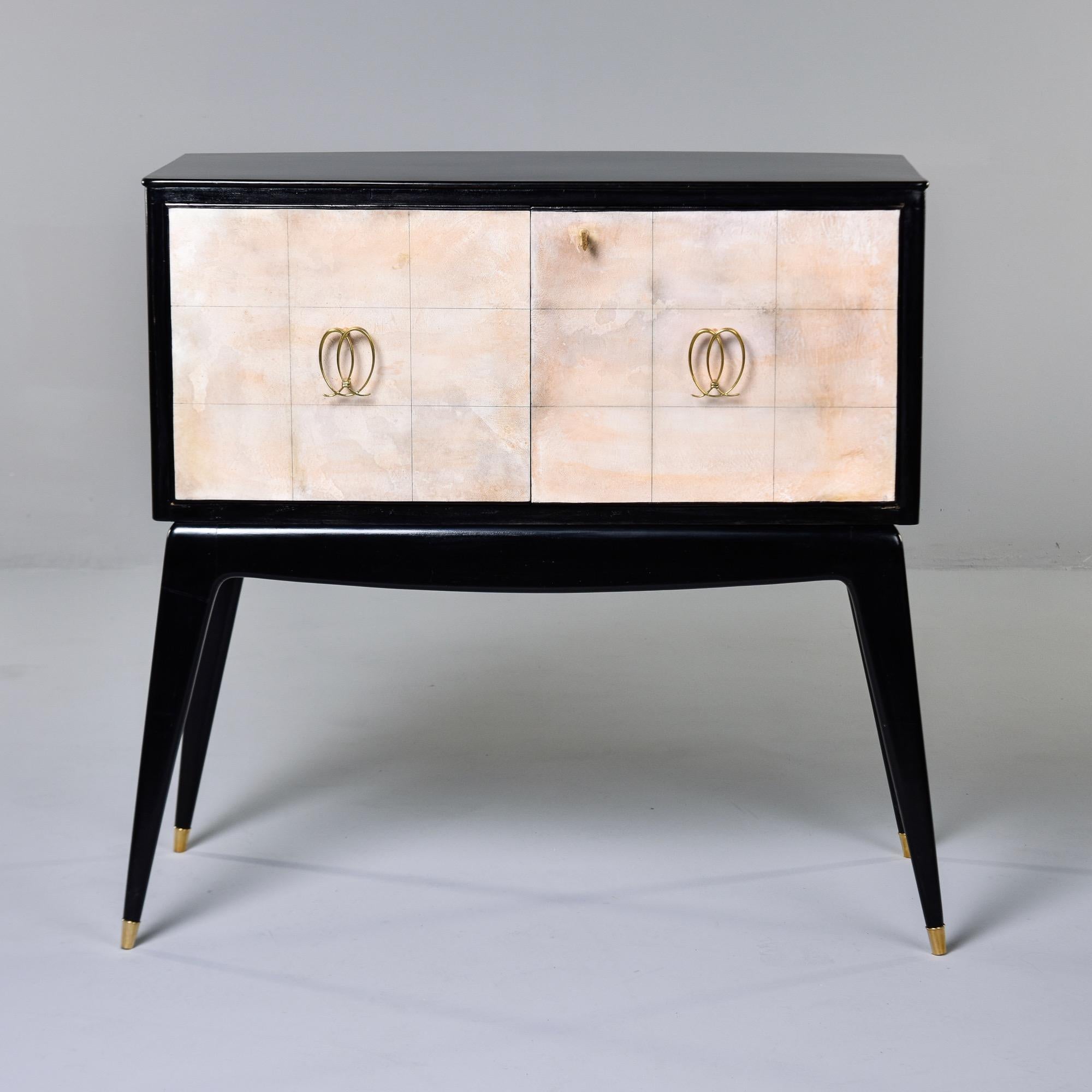 Found in Italy, this mid century Italian bar cabinet has an ebonised body, parchment covered doors with a functional lock and a mirrored interior. Cabinet has original brass hardware, internal bottle racks, checkered mirrored sides, and slim tapered