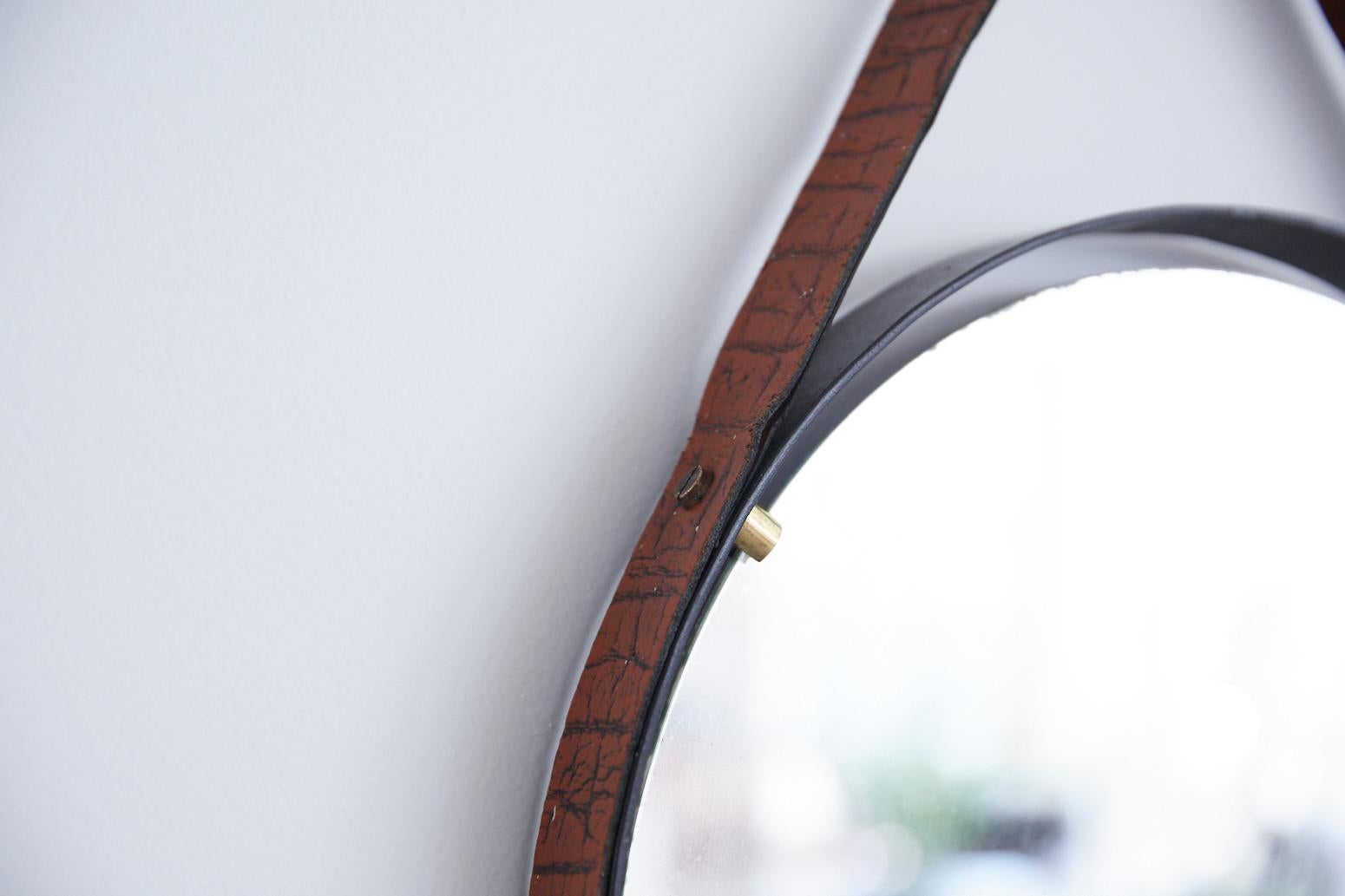 Beautiful circular Italian floating mirror from the 1970s. Round black iron frame holds floating mirror with brass pegs. Iron frame wrapped in textured brown leather with strap and metal stud detailing. Leather with slight cracking and mirror has