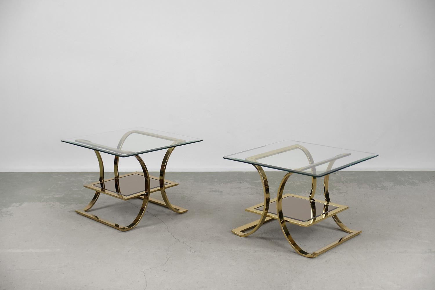 This pair of glass tables was produced in Italy during the 1960s. The upper glass is transparent and the lower glass is mirrored. The base is made of a hand-bent gold-colored metal. The tables are slightly different from each other in size. This set