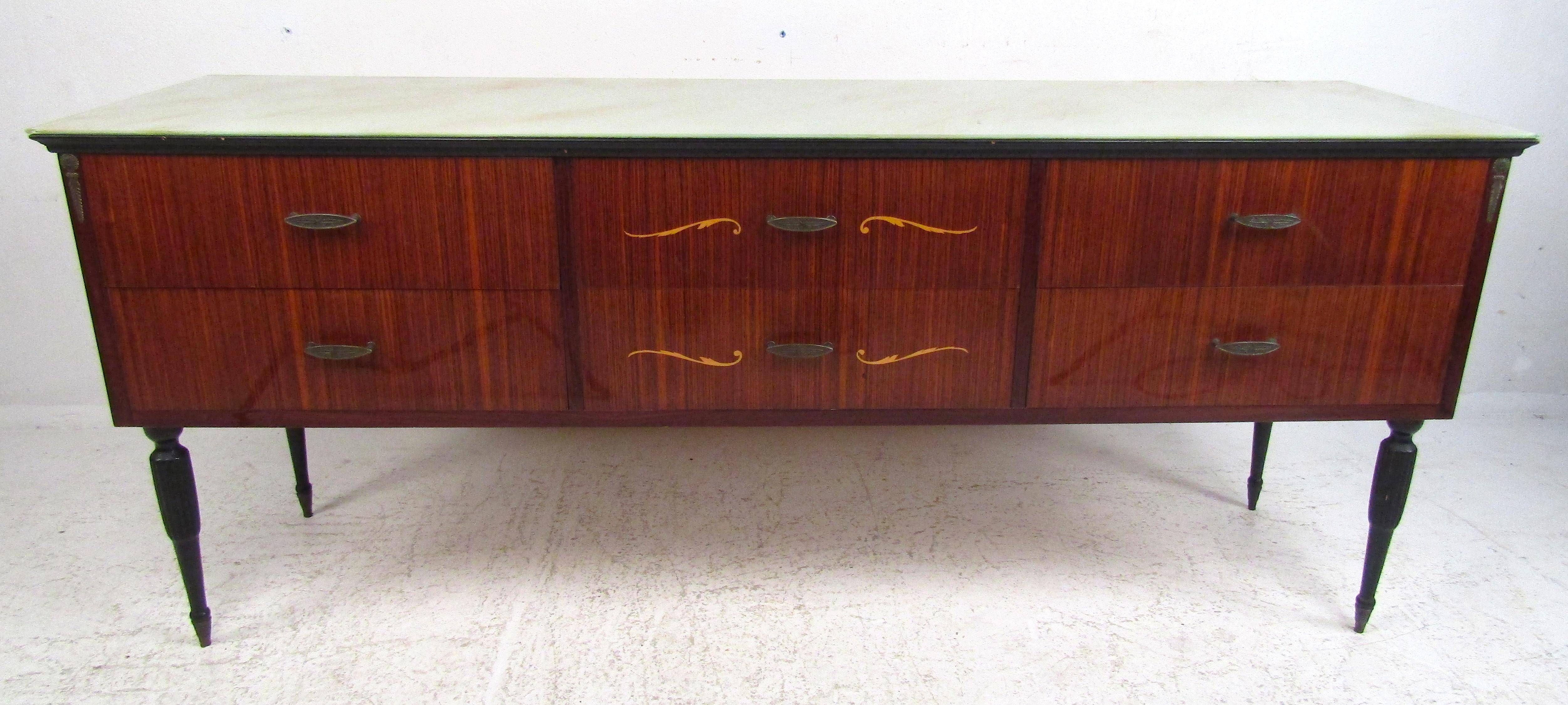 A fabulous midcentury Italian dresser that boasts an elaborate glass top, carved legs, and unique inlays on the drawer fronts. This well-made case piece shows attention to details with its brass oval drawer pulls and unusual decorative fixtures on