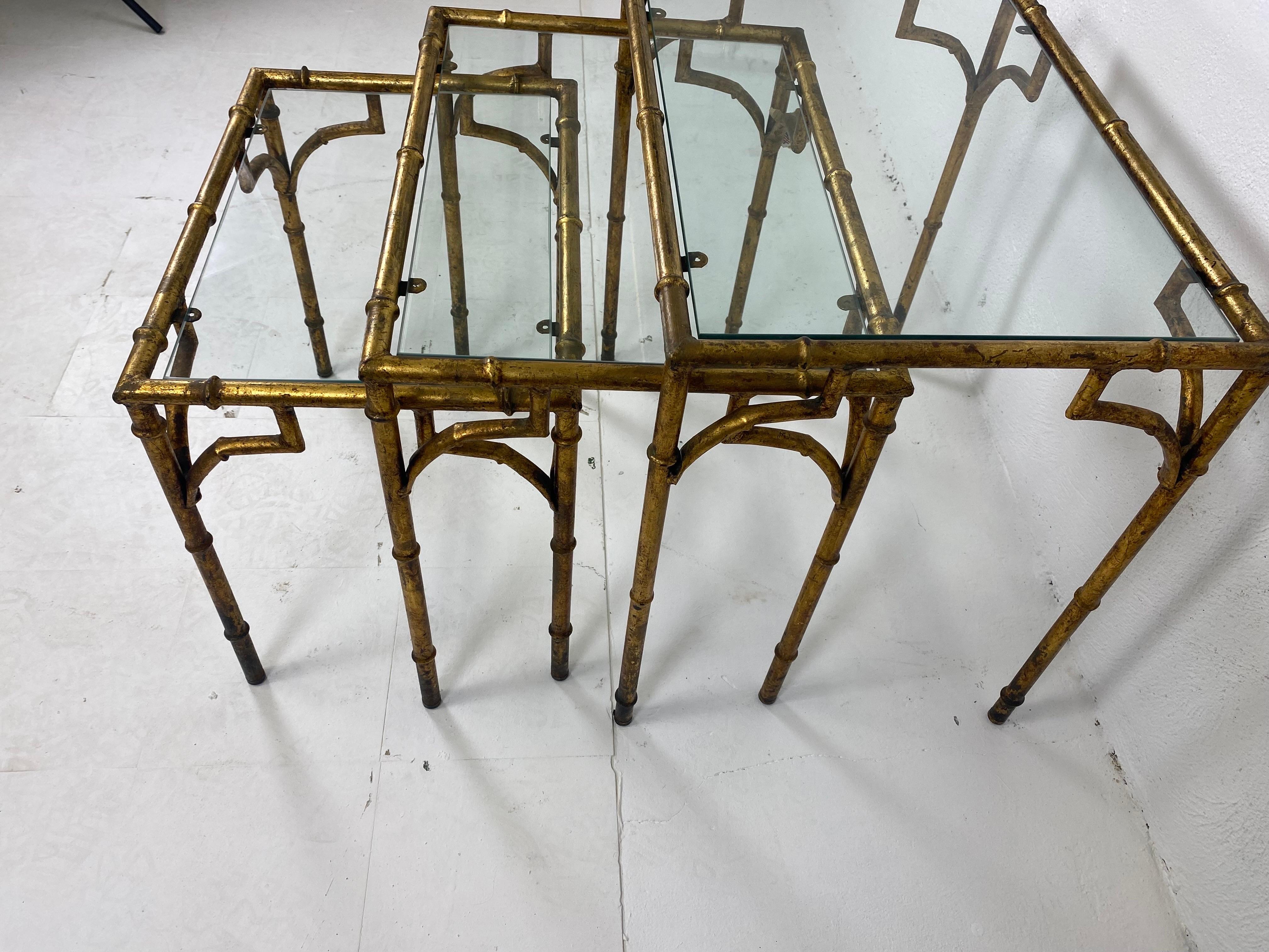 This is a vintage set of Italian gold gilded wrought iron faux bamboo nest of tables. Each table has a glass top and the tables snuggly fit together underneath the larger table. The gold leafing has a distressed finish to the surface. These tables