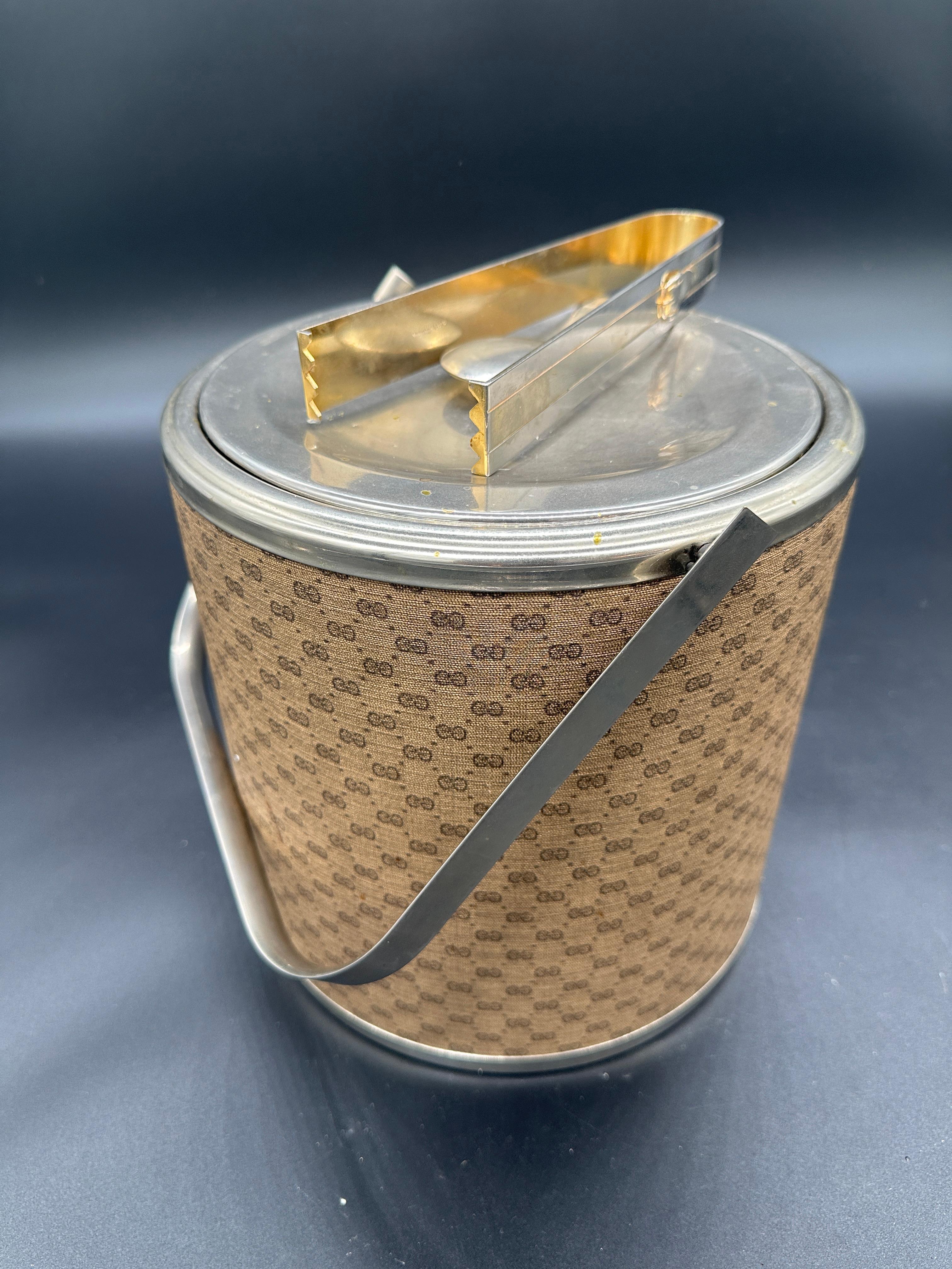 This vintage Gucci ice bucket from the 1980s features the iconic GG monogram pattern, a sturdy handle, and tongs, embodying classic Italian luxury and style.

