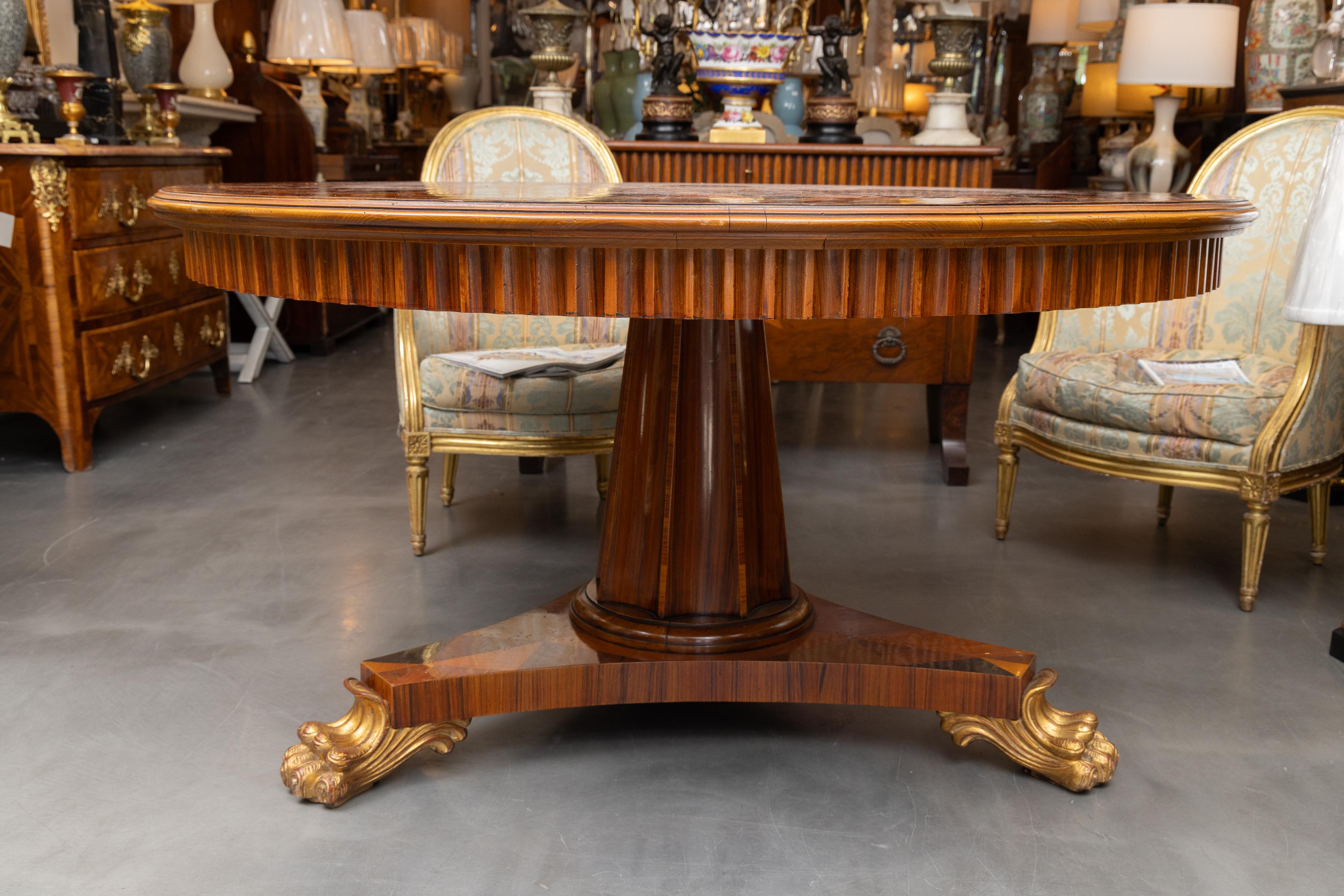 Se trata de una singular mesa de centro italiana de época, con el tablero de parquet de varias maderas sobre un prominente friso acanalado y pedestal, situado sobre una base tripartita que termina en tres patas doradas.