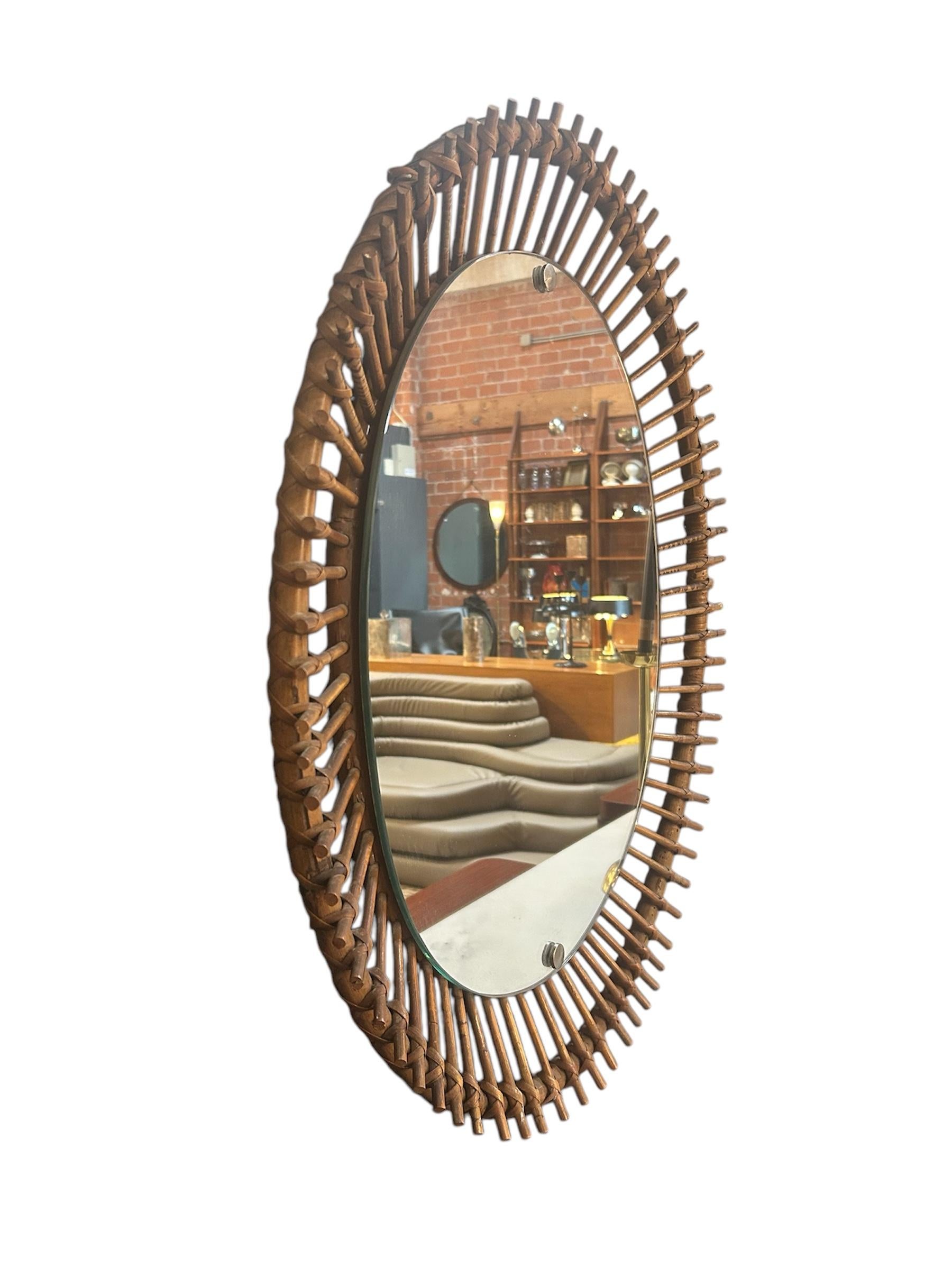 Un affascinante specchio da parete vintage italiano degli anni '60, caratterizzato da un vetro ovale centrale incastonato in una scultorea cornice in rattan intrecciato a mano. Il motivo radiale evoca un effetto a raggiera, fondendo il calore