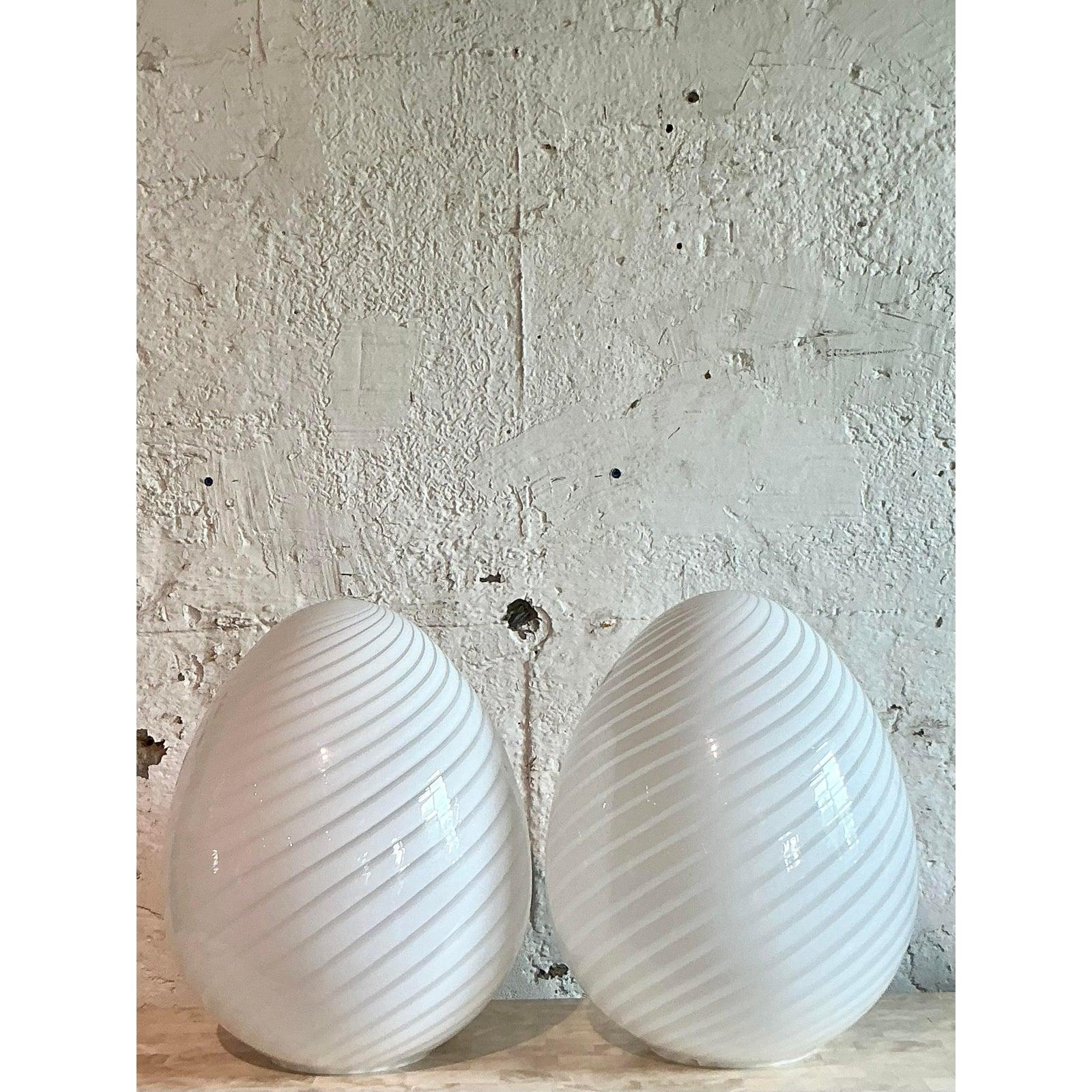 Proyecta un cálido y etéreo resplandor en tu espacio con este impresionante par de lámparas de huevo de cristal remolinado italiano vintage. Fabricada en el célebre estilo de Murano, cada lámpara muestra delicadas cintas de cristal blanco que se
